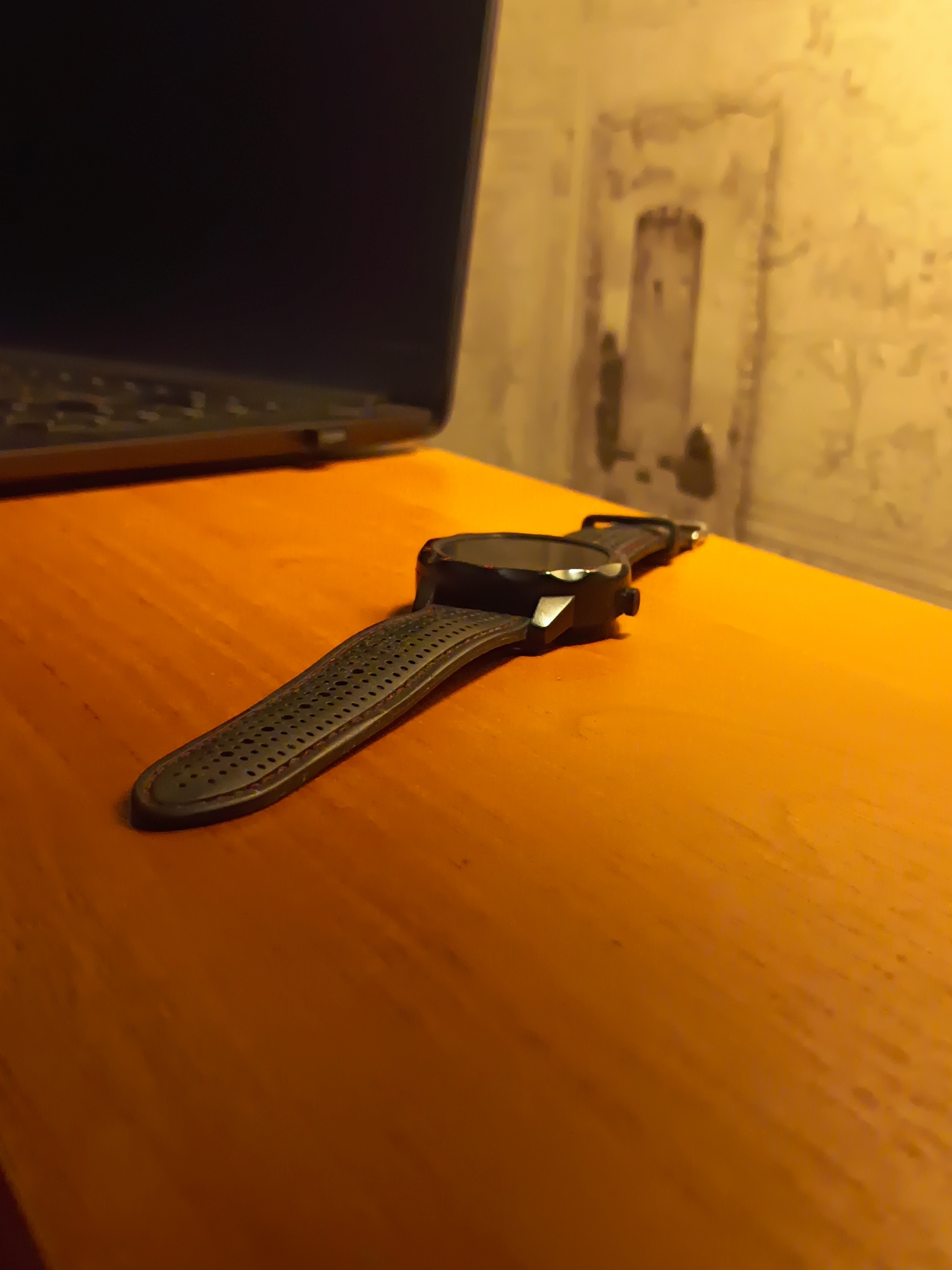 A close-up, low-angle shot shows a black digital watch with a perforated strap lying on an orange wooden surface. The watch is angled from left to right, with its face out of focus in the background. To the left, a portion of a dark laptop is visible. In the background, a textured, light-colored wall with some dark markings is out of focus. The lighting is warm, suggesting an indoor setting, possibly a home or office. The image does not provide any information that would allow for the determination of its location in the city of Izmail, Ukraine.