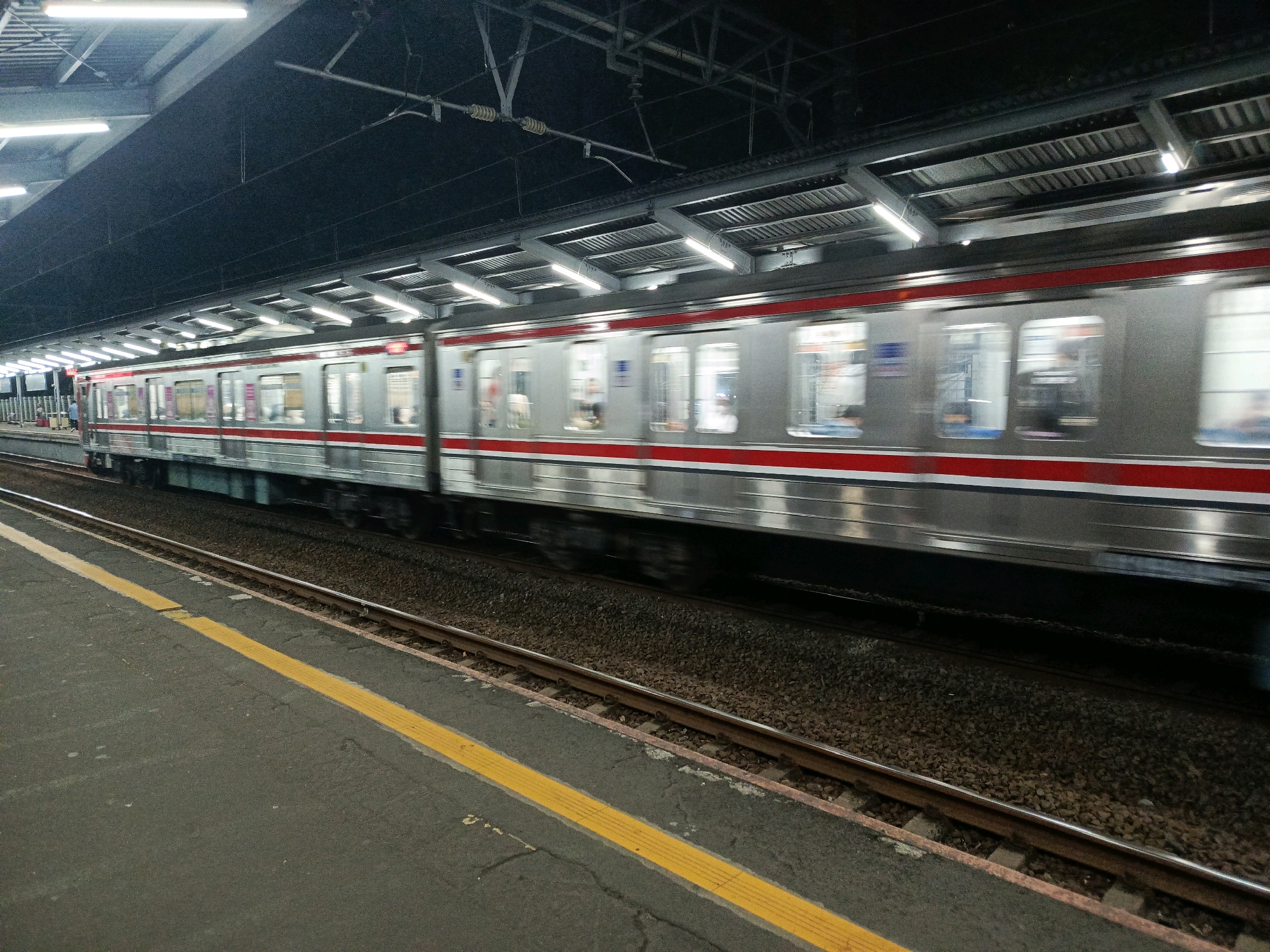 A train is speeding past a station platform. The train is silver with a red stripe and has many windows. The platform is paved with asphalt and has a yellow safety line. The station appears to be covered by a roof supported by metal beams, and there are lights on the ceiling. The train tracks are visible, covered with gravel. It is unclear if this scene is in South Jakarta, Indonesia, as there are no identifying markers.