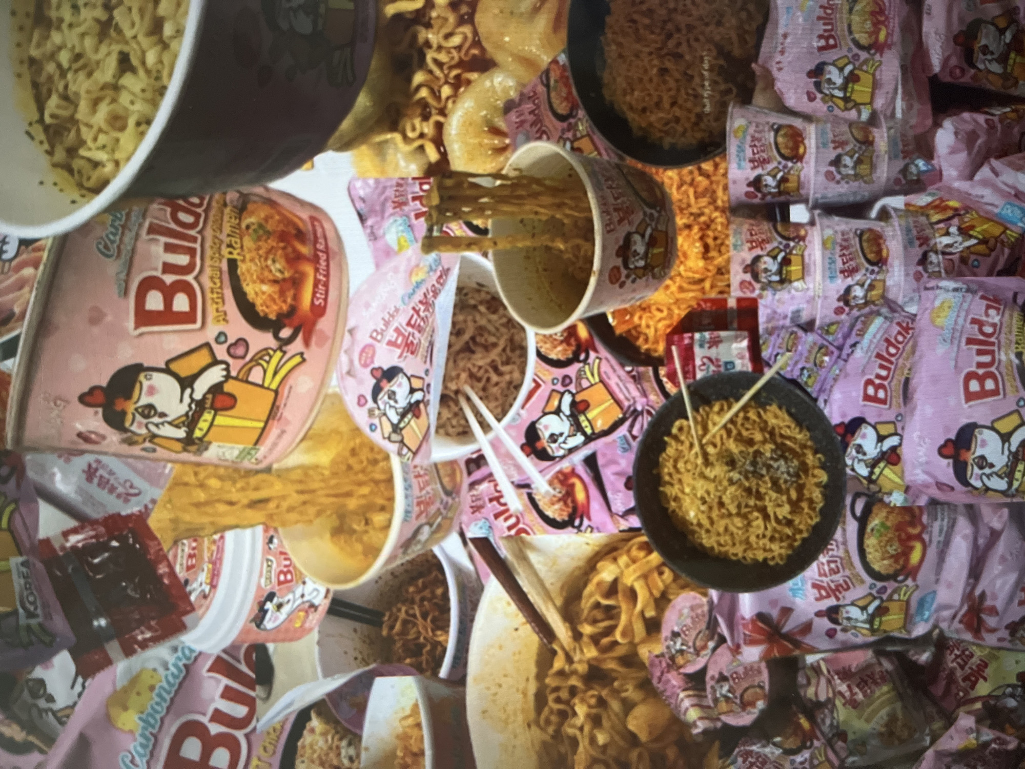 The image is a collage of instant ramen products, primarily featuring the Buldak brand. The dominant colors are pink and yellow, with a strong emphasis on the cartoon mascot of the brand. Various forms of the ramen are visible, including cups and packets, some of which appear to be cooked and ready to eat, with noodles and broth. The products are arranged in a cluttered, disorganized manner, suggesting a display or a collection of food items. The background is a mix of other ramen packaging and a blurry, abstract pattern. There are no people or discernible environmental features of a specific city or location.
