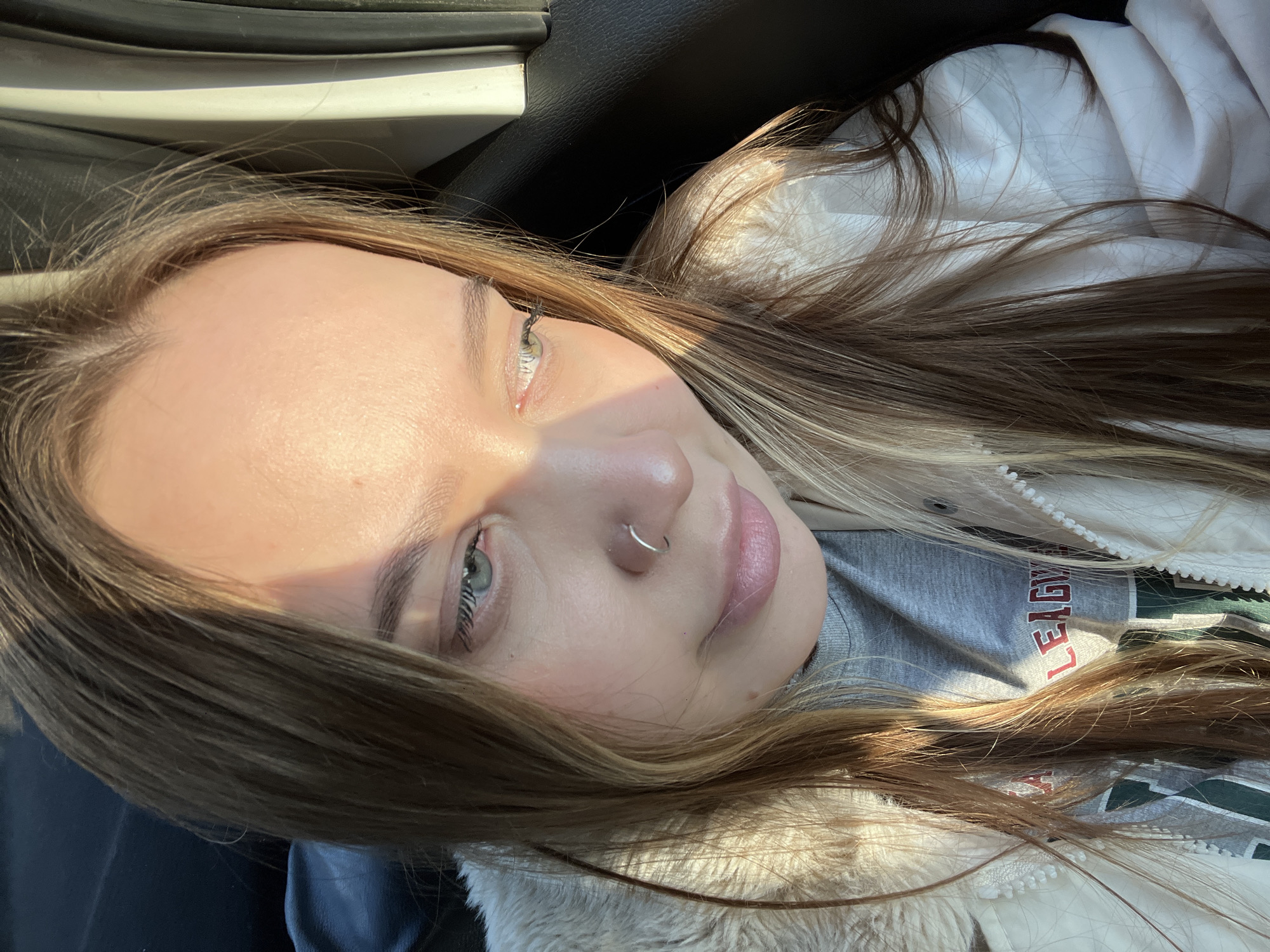 A young woman with light skin and green eyes is facing the camera at an angle. She has long brown hair with blonde highlights, a silver nose ring, and light pink lips. She is wearing a white jacket with fur trim and a gray sweatshirt with red lettering. The background includes a dark interior, possibly a car, and sunlight is shining on her face.