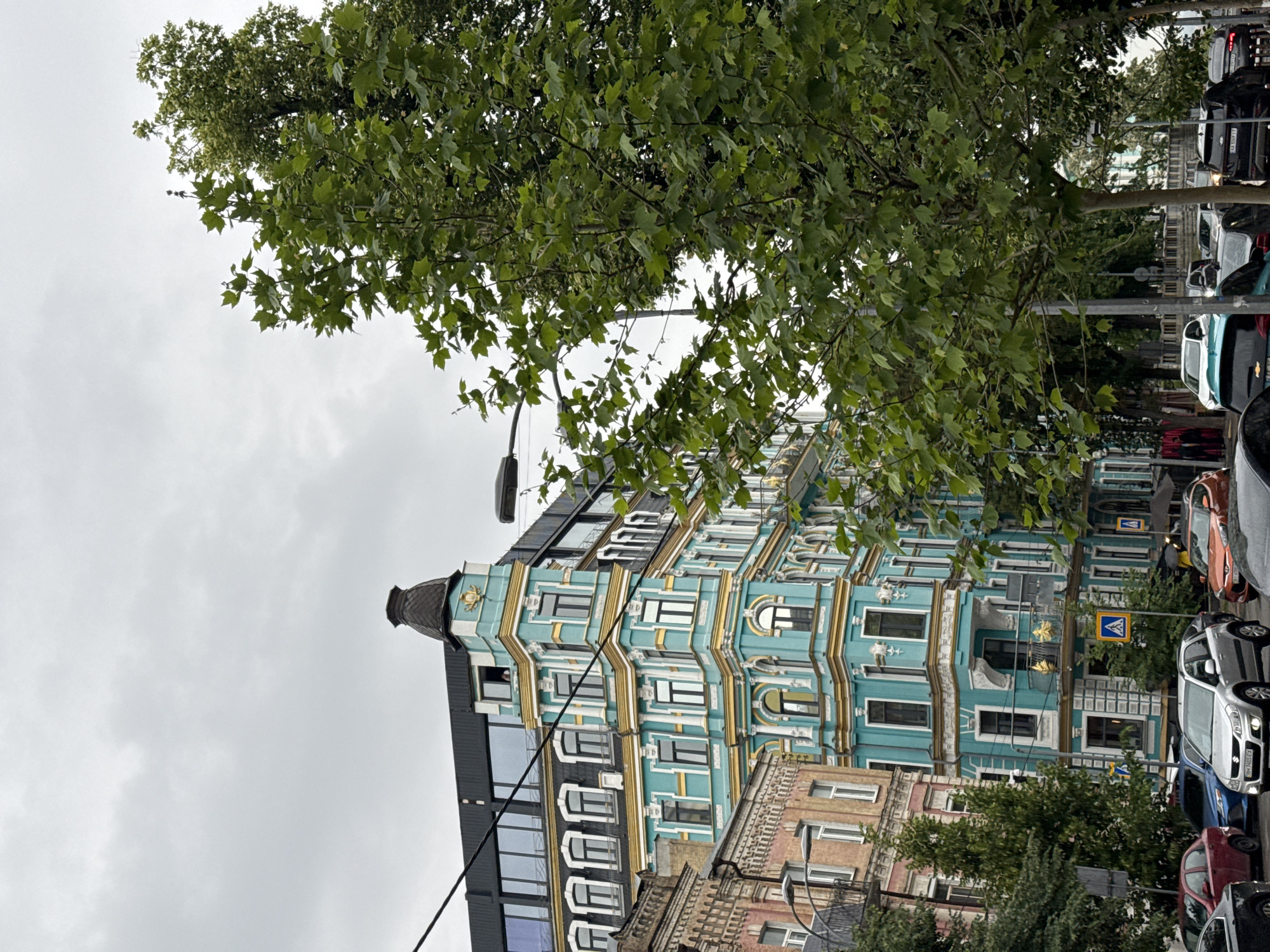 This image captures a vibrant urban street scene in Kyiv, Ukraine, characterized by striking architecture and lush greenery under an overcast sky.
The central subject is a multi-story building featuring an ornate facade painted in a distinctive turquoise or teal color, beautifully contrasted with golden-yellow decorative accents and white window frames. The building exhibits a mix of architectural styles, with some sections having classically inspired arched windows and intricate detailing, while others incorporate more modern, dark glass panels. A dark, conical turret or cap crowns the upper-left corner of this prominent structure.
Abundant green trees, likely plane trees with their characteristic lobed leaves, occupy the foreground and left side of the frame, partially obscuring the sky and upper sections of the buildings. The sky is uniformly light grey and cloudy, suggesting an overcast day.
On the right side of the image, a street is visible, lined with numerous parked cars of various models and colors, including silver, black, orange, and red. Visible text includes "SUZUKI" on the rear of a silver car and a red car. License plates are partially visible, with "AA 0340 EY" discernible on a grey car and "AA 0300" (or similar) on a black car. A blue square pedestrian crossing sign with a white walking figure is also present near the parked vehicles. Portions of other, less ornate buildings with brick-colored facades are visible in the lower right, adding to the urban density of the scene. The overall impression is that of a bustling city street during daylight hours.
