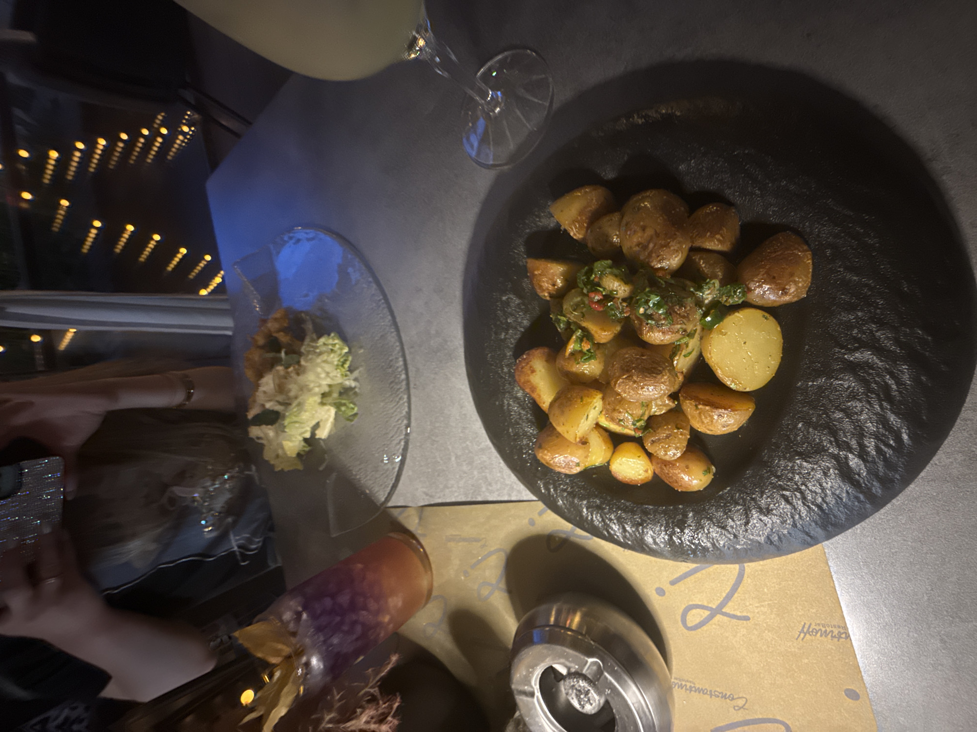 The image shows a table setting in what appears to be a restaurant. The scene includes a plate of roasted potatoes with herbs on a black plate, a small plate of what looks like a cabbage salad, and a glass of a pale-colored drink, possibly lemonade or juice. There is also a decorative vase with dried plants and an ashtray on the table. The table covering has writing that reads “Constantinoff Restaurant”. Based on this, the location is Sofia, Bulgaria, as Constantinoff Restaurant is a known establishment in the city. A portion of a person is visible holding a phone and is taking a photo of the food. The background shows a window with multiple light dots, suggesting an urban environment at night. The overall atmosphere seems to be a relaxed dining experience.