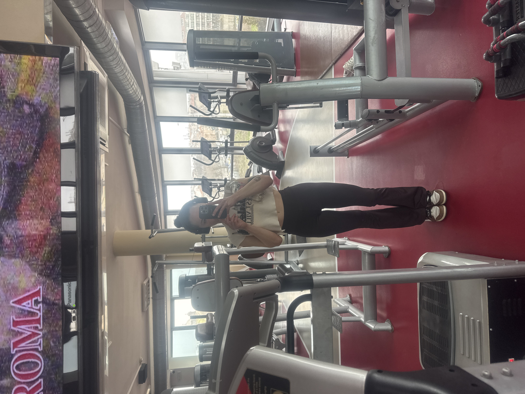 The image depicts a female individual taking a selfie in a large wall-mounted mirror within a fitness center. The woman is positioned centrally, wearing a white short-sleeved crop top with a dark cityscape graphic, dark flared athletic pants, and brown and white sneakers. Her hair is styled in a bun, and she is wearing dark headphones. Tattoos are visible on her right forearm and left bicep. She holds a dark-colored smartphone, directed towards her reflection.
The fitness center features a predominantly red floor with some grey sections. Various pieces of exercise equipment are visible, including the partial reflection of a silver and black cardio machine in the foreground on the left, a grey strength training machine with red padding behind the subject, and several elliptical trainers and stationary bikes further back near the windows. A pair of white sneakers rests on the floor near equipment to the right of the subject's feet.
The facility has large, floor-to-ceiling windows that offer an expansive view of an urban landscape. Through these windows, multiple residential and commercial buildings are visible, along with trees and what appears to be a distinct Orthodox church with a prominent dome and cross. This church is identifiable as the Saints Cyril and Methodius Cathedral, confirming the location as the city of Burgas, Bulgaria.
Above the windows, a white ceiling features exposed silver ventilation ducts and artificial lighting fixtures. A large flat-screen television is mounted high on the wall, displaying content with the word "ROMA" clearly visible, along with a "Panasonic" logo below it. A light-colored structural pillar is also present in the room.