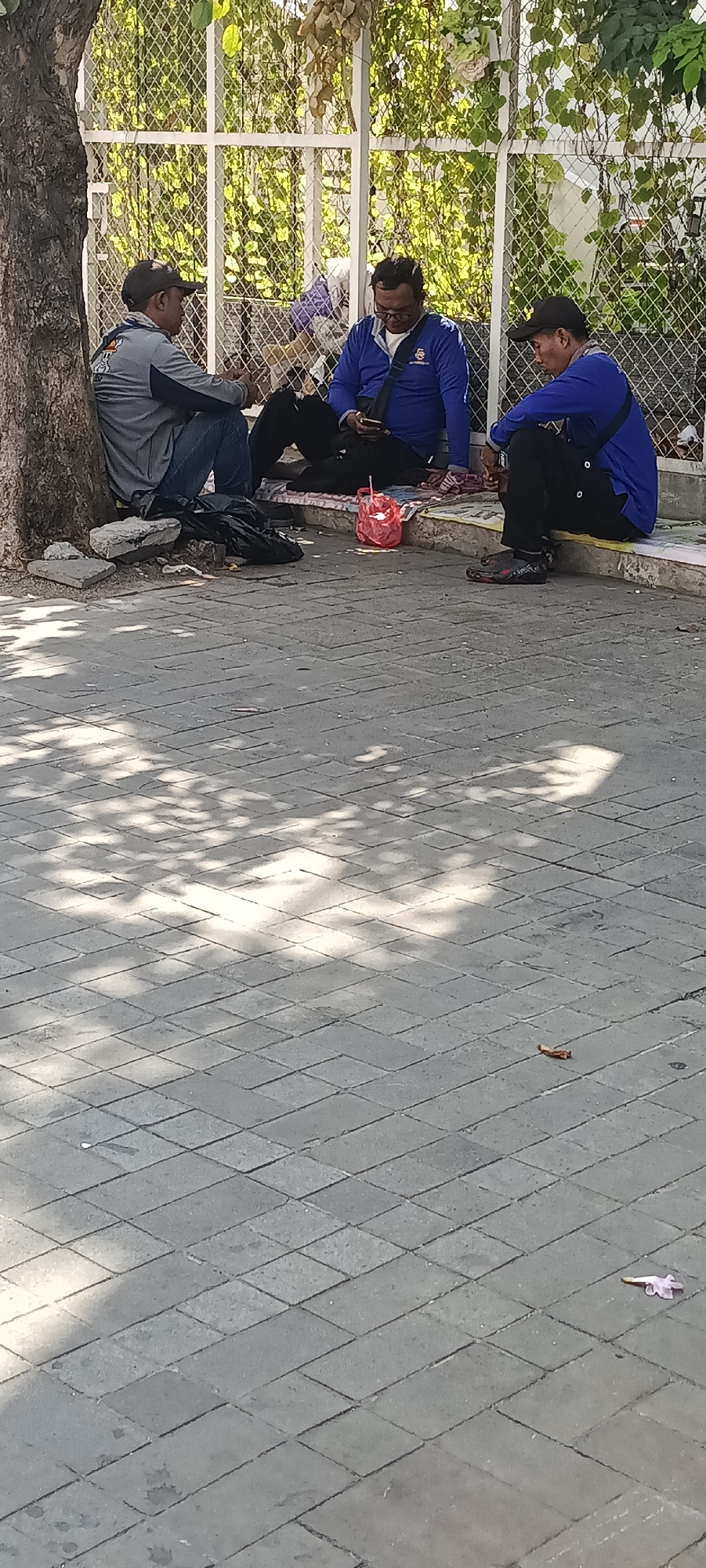 Three men are seated on a paved walkway in front of a white fence with climbing vines. The man on the left is wearing a gray long-sleeved shirt, blue jeans, and a black baseball cap. He is leaning against a tree trunk. The middle man is wearing a blue long-sleeved shirt, black pants, and glasses. He is looking at his phone. The man on the right is wearing a blue long-sleeved shirt and black pants. He is also looking at his phone. A red plastic bag is placed on the ground between the middle and right men. There is a black trash bag near the man on the left. The walkway is paved with rectangular stones arranged in a diamond pattern. Sunlight casts shadows from the tree and vines onto the walkway. The background suggests an outdoor urban environment, possibly near a park or public space in Semarang, Indonesia.