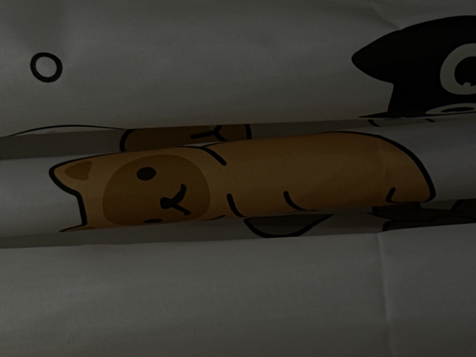 The image shows a close-up of a white fabric curtain with cartoon cat patterns. One cat is yellow with a black nose and eyes, wearing a brown costume with a black outline. Another cat is black with white eyes and is partially visible on the right side. The curtain is rolled or folded, creating a shadow and highlighting the texture of the fabric. There is a dark circular shape on a white background on the upper right of the image, possibly part of another cat's face or a design element. The image is dimly lit, with soft shadows. The location is the city of Poritsy, Russia.