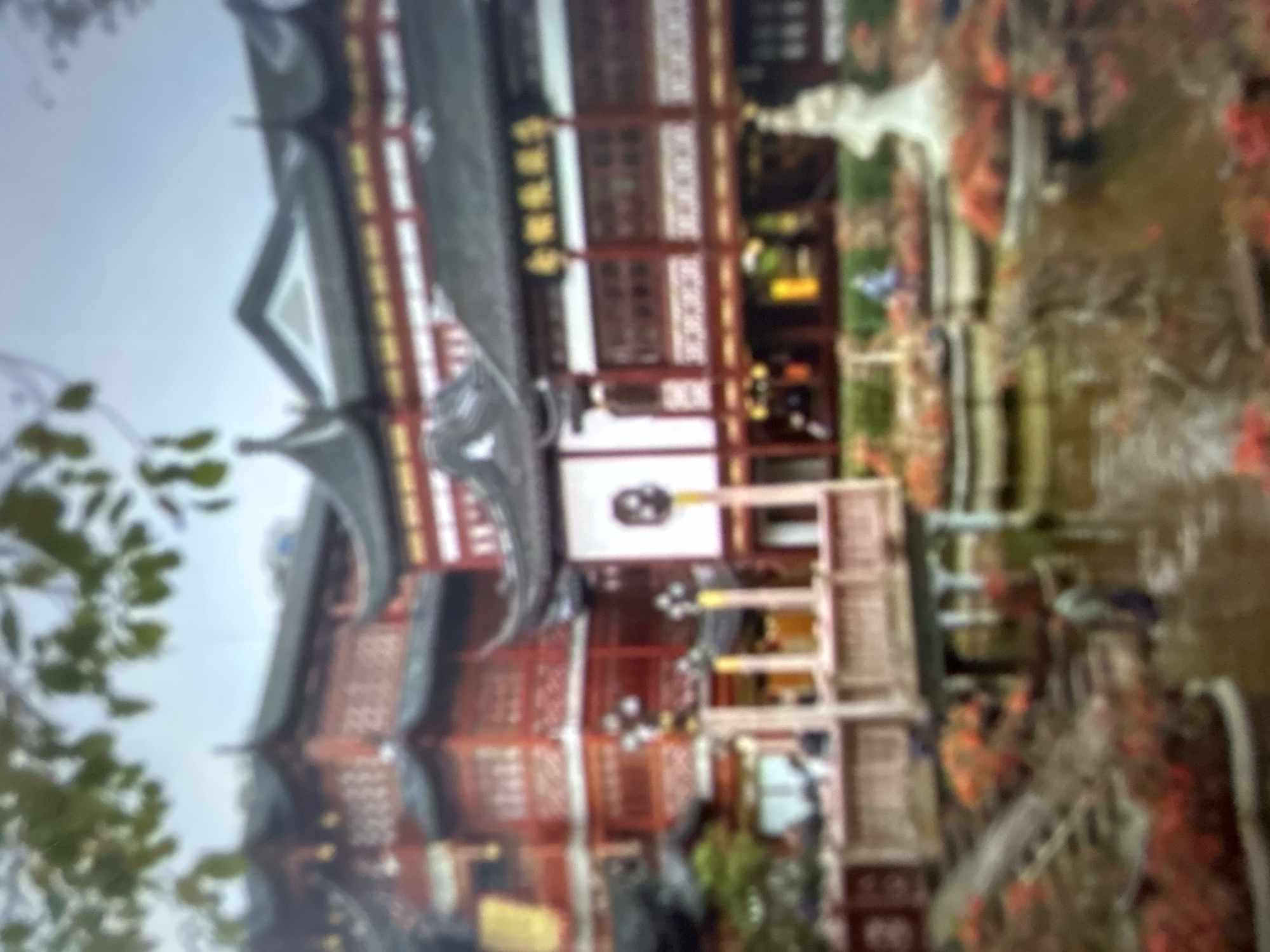The image depicts a traditional Asian temple or pavilion, characterized by its multi-tiered, dark tiled roof with upturned eaves, and red wooden structures adorned with intricate white latticework. The main building features multiple windows and what appears to be decorative elements. To the right of the building, a body of water, possibly a pond or stream, is visible, with reflections of the surrounding environment. Lush greenery, including trees and foliage, frames the scene, suggesting an outdoor setting. The presence of flowers, some of which are reddish-orange, adds a touch of color and vibrancy to the landscape. The overall atmosphere of the image is serene and picturesque. The resolution is blurry, making it difficult to discern specific details like people or their activities. There are some blurry characters visible on a vertical sign on the building, but they are illegible. The image appears to be taken during daylight, and the lighting suggests it might be an overcast or slightly hazy day. Based on the provided context, the location is Gelendzhik, Russia, but the architectural style strongly suggests an East Asian influence, creating a juxtaposition with the given location.