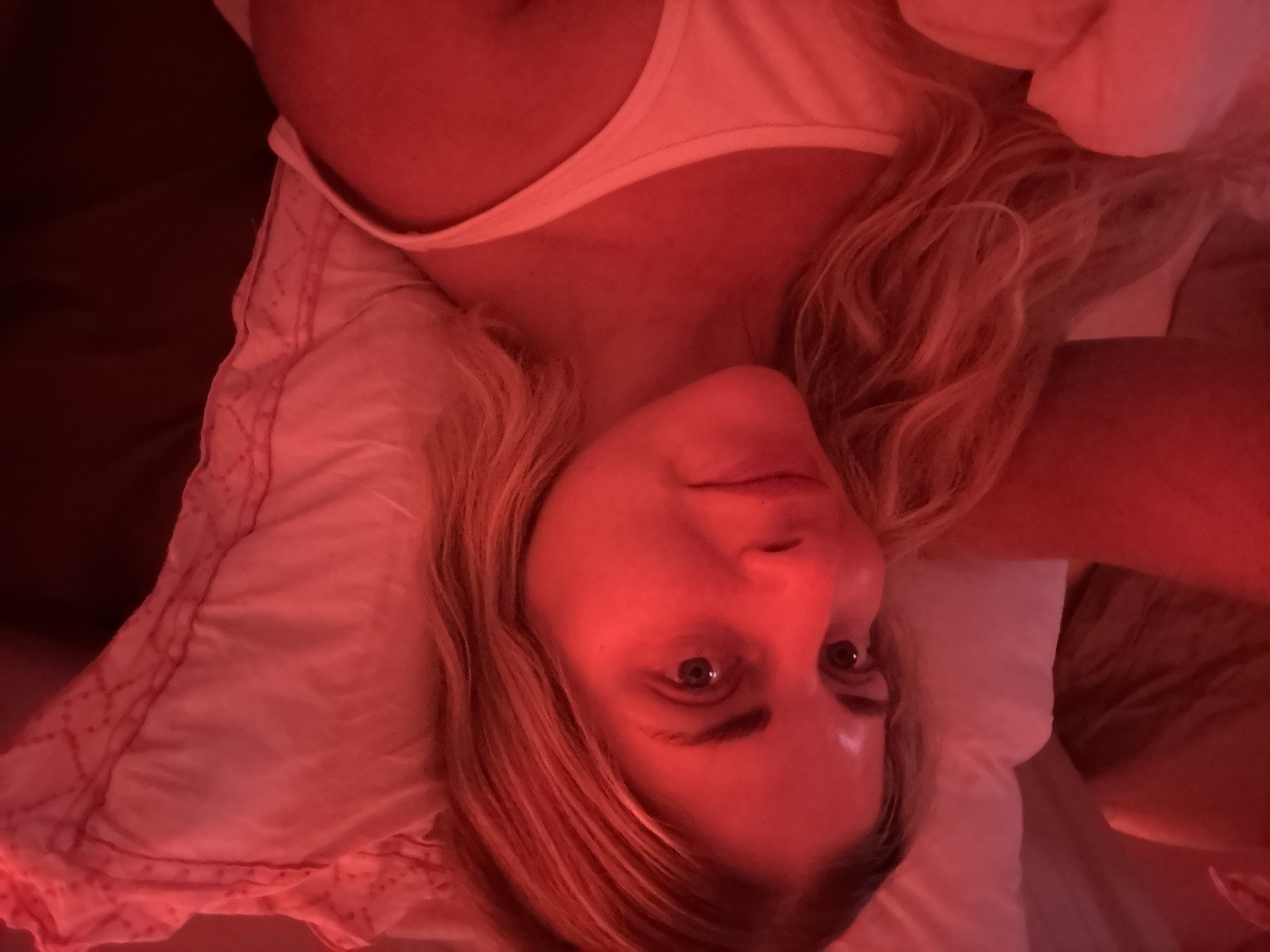 This is a close-up selfie of a woman in bed, lit by red light. She is lying on her side, with her head resting on a pillow. Her hair is blonde and wavy, and she is wearing a white tank top. The background is a bed with white sheets and a pink floral patterned pillowcase. The image does not provide any information about the location in Paris, France.