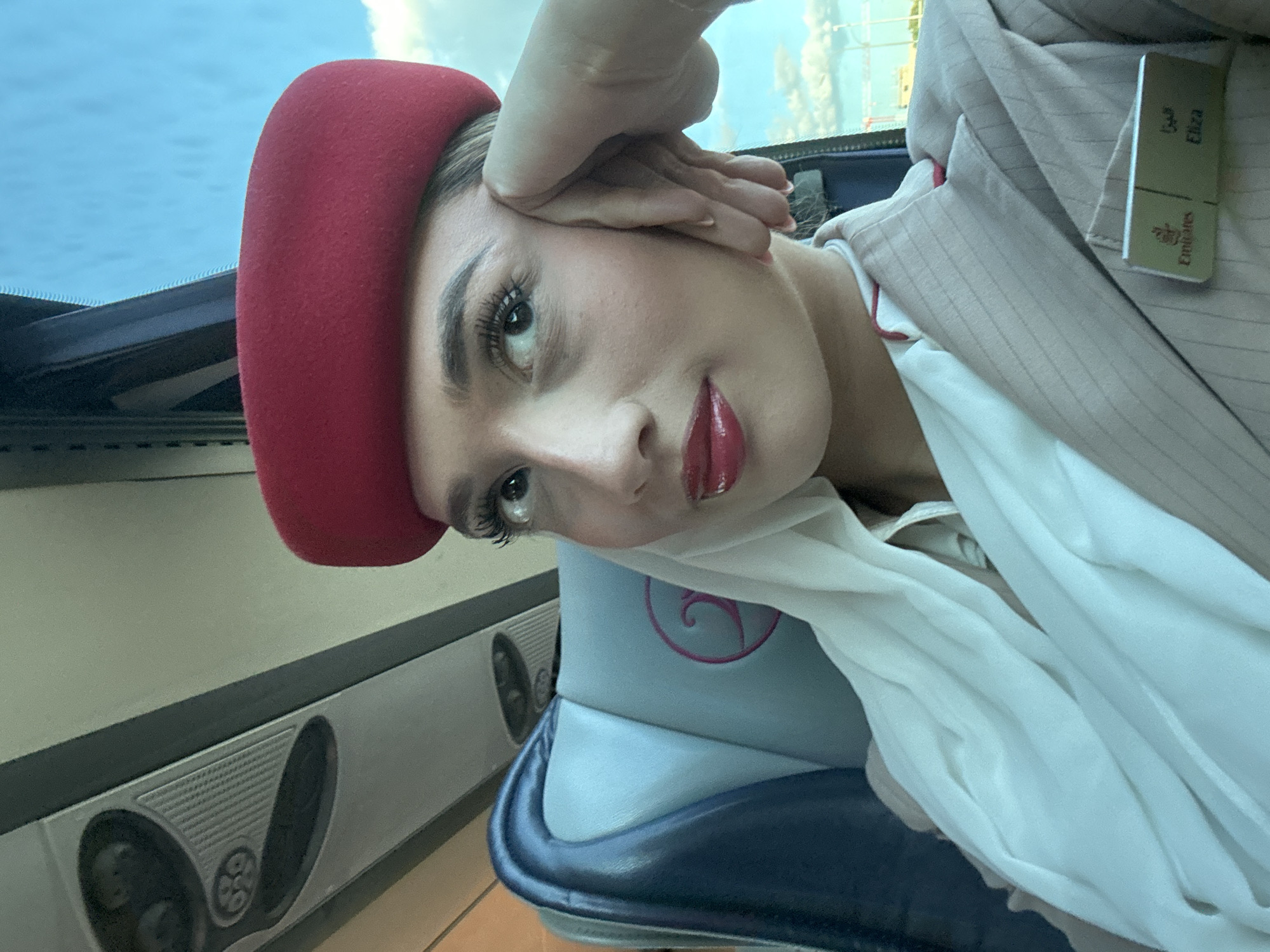 A woman, likely a flight attendant based on her uniform including a red hat and Emirates Airlines name tag, is sitting in what appears to be an airplane seat near a window. Her hand is resting on her forehead. The location is unlikely to be in Madrid, Spain, because the image depicts the interior of an airplane.