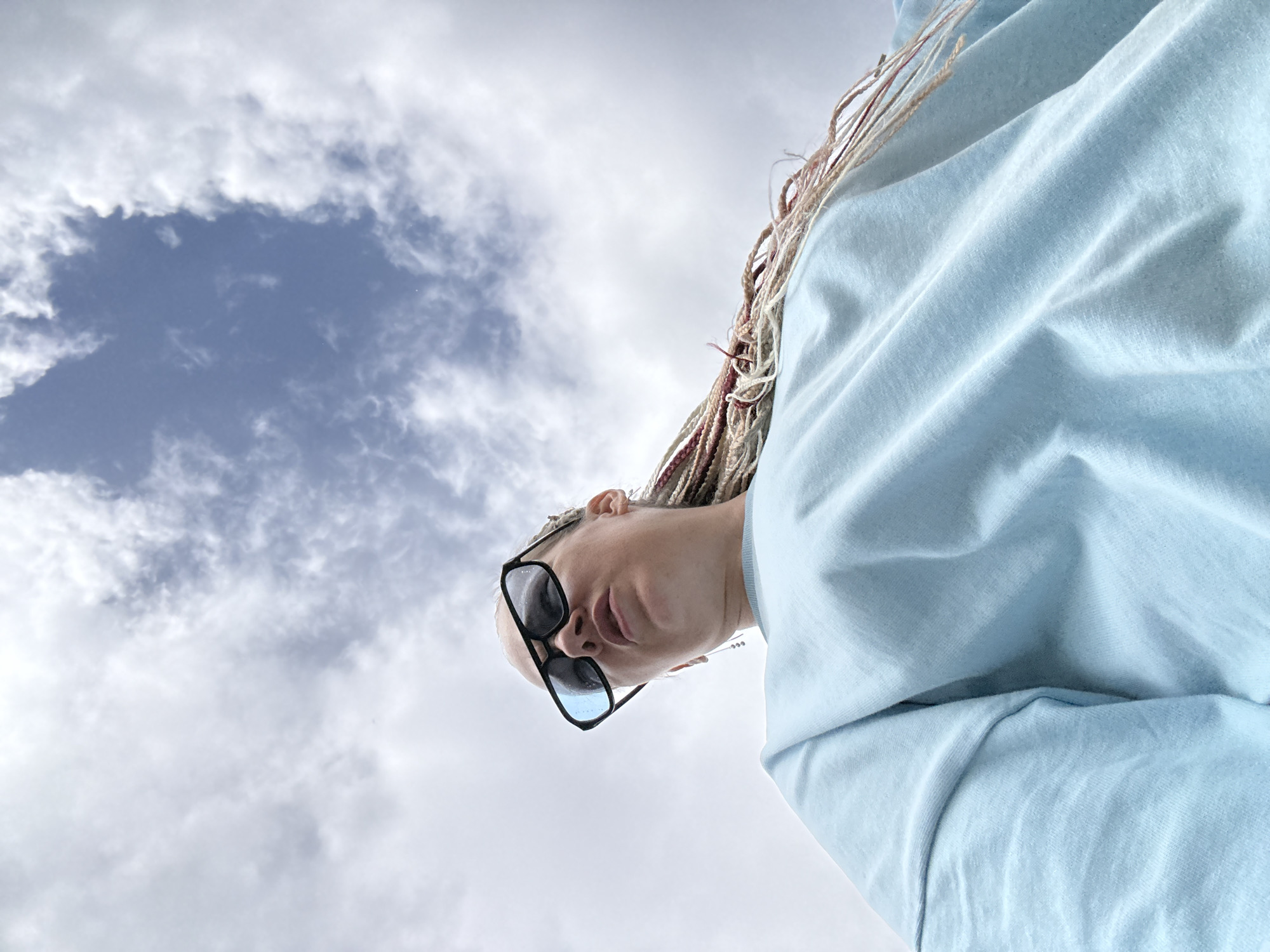 The image is a low-angle, full-body shot of a woman wearing a light blue sweatshirt and large black sunglasses. She has blonde hair styled in many thin braids, some of which are pink and blonde. She is looking upwards towards the sky, which is partly cloudy with patches of blue visible. The woman's facial expression is difficult to determine clearly due to the angle and the sunglasses. The image is taken outdoors in an urban environment, likely a city. There is no specific information to pinpoint the location to Moscow, Russia.
