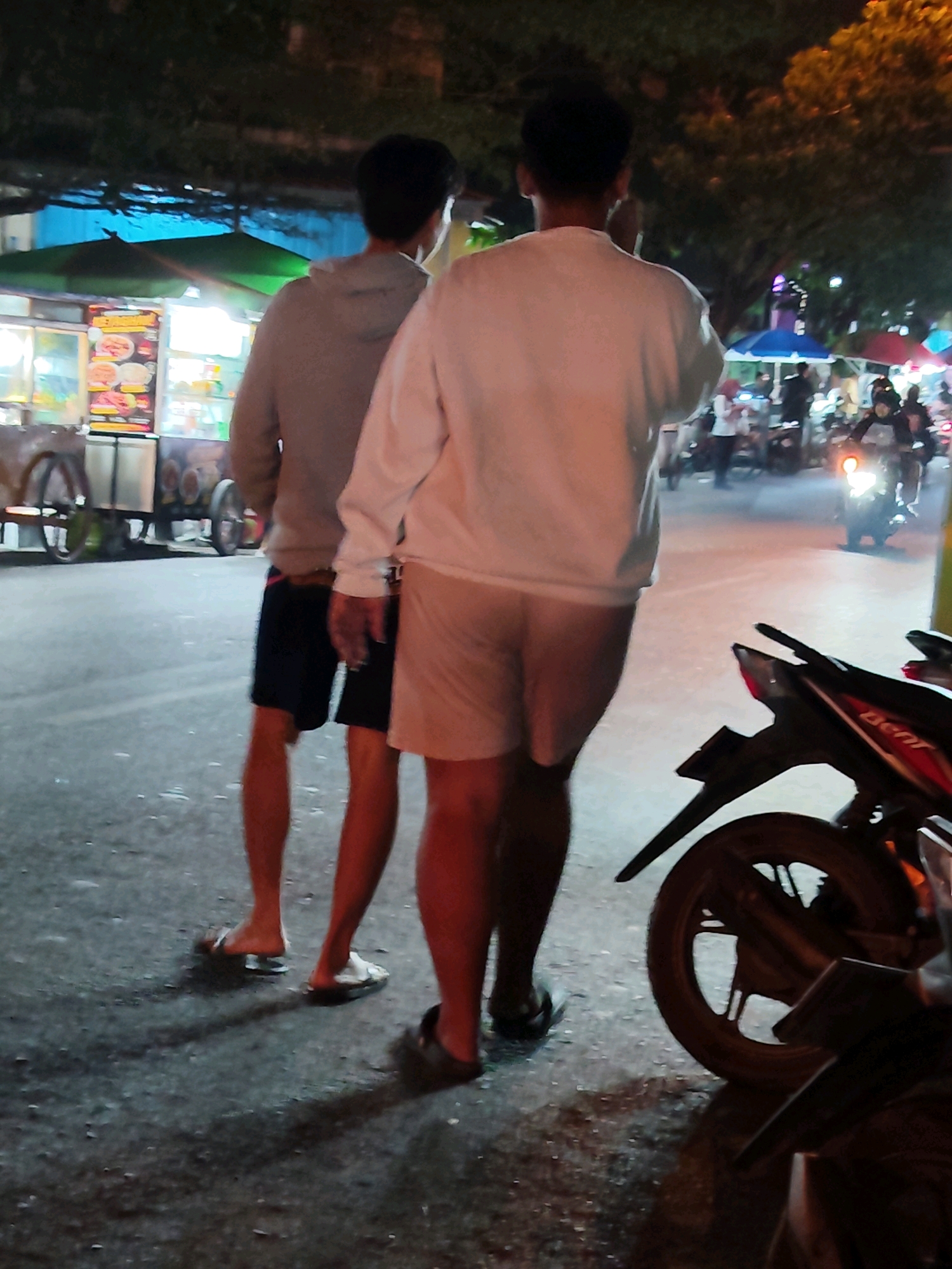 The image depicts an outdoor scene at night, likely in Purwakarta, Indonesia, possibly a street market or event. Two individuals are standing in the foreground, facing away from the camera. The person on the left wears a grey hoodie and dark shorts. The person on the right is wearing a white long-sleeved shirt and light-colored shorts.
In the background, there are several stalls or vendors, one with bright lights and display boards featuring food items. Various motorbikes are visible, some ridden by people, and a motorcycle is parked in the right foreground. The ground appears wet, suggesting recent rain. The overall atmosphere suggests a lively, perhaps crowded, night-time urban environment.
