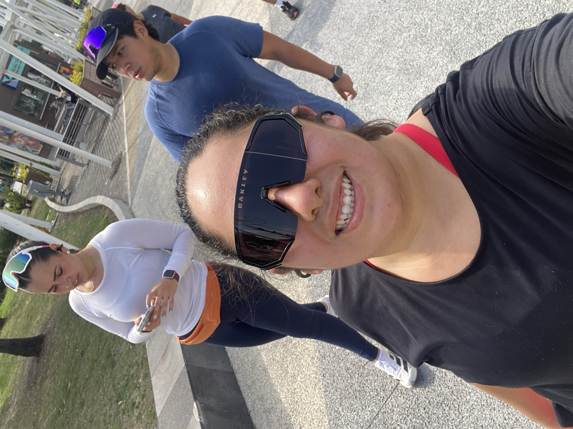 A selfie captures a group of four people on a sunny day, likely in a park. The person taking the photo is wearing Oakley sunglasses, a black tank top over a red sports bra, and their hair is pulled back. To their left, a person in a white long-sleeved shirt, blue leggings, an orange fanny pack, and white sneakers is holding a cell phone. Behind the selfie-taker and to the left is a person in a blue shirt. A fourth person, only partially visible, is further back. The background includes trees, grass, a grey pavement, a modern structure with vertical bars, and a building with a sign in Spanish. Based on the architecture and signage, this location is likely in the city of Puebla, Mexico.