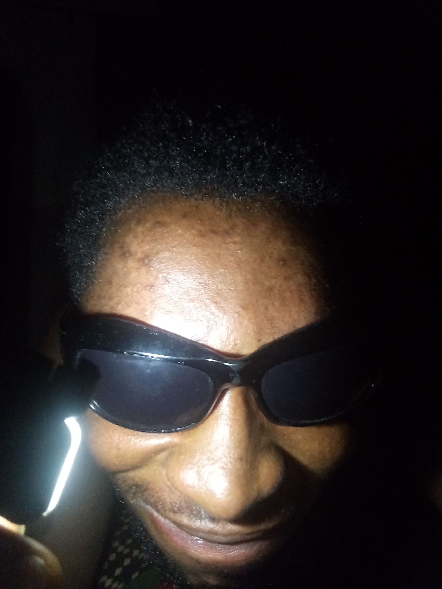 A person with dark skin is wearing sunglasses and facing the camera. A light source is visible on the left side of the frame, illuminating the face partially. The background is dark and obscured, making it difficult to discern any specific environmental details. It is not possible to determine the location within Lokoja, Nigeria, or any specific actions or interactions from the provided image.
