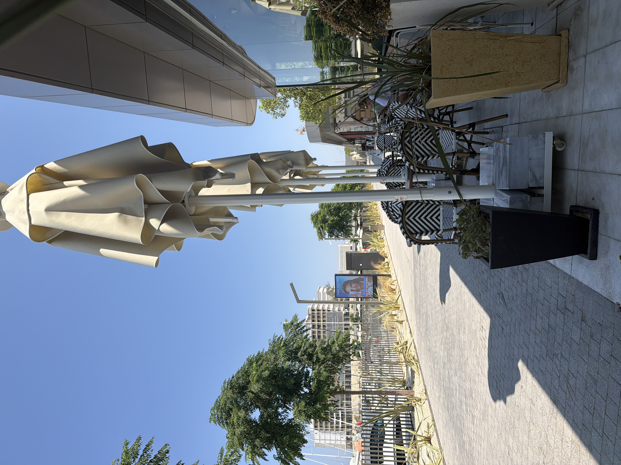 The image depicts a sunny day at a waterfront establishment in Kuşadası, Turkey, likely a cafe or restaurant. Several large, closed cream-colored umbrellas are positioned on a paved walkway. To the right, a modern building with reflective windows is visible. In front of the building, there are tables and chairs with black and white patterned upholstery. A person is seated at one of the tables, appearing to be reading. To the left of the umbrellas, a paved pathway lines a metal fence, beyond which are trees, buildings, and boats in a harbor, indicative of a marina setting. A digital billboard displays an advertisement featuring a woman's face and text, including "Yves Rocher" and "50 SPF BB KREM." The ground is paved with rectangular stones, and planters with greenery are placed strategically. The overall scene is one of an outdoor seating area adjacent to a commercial building in a coastal city.