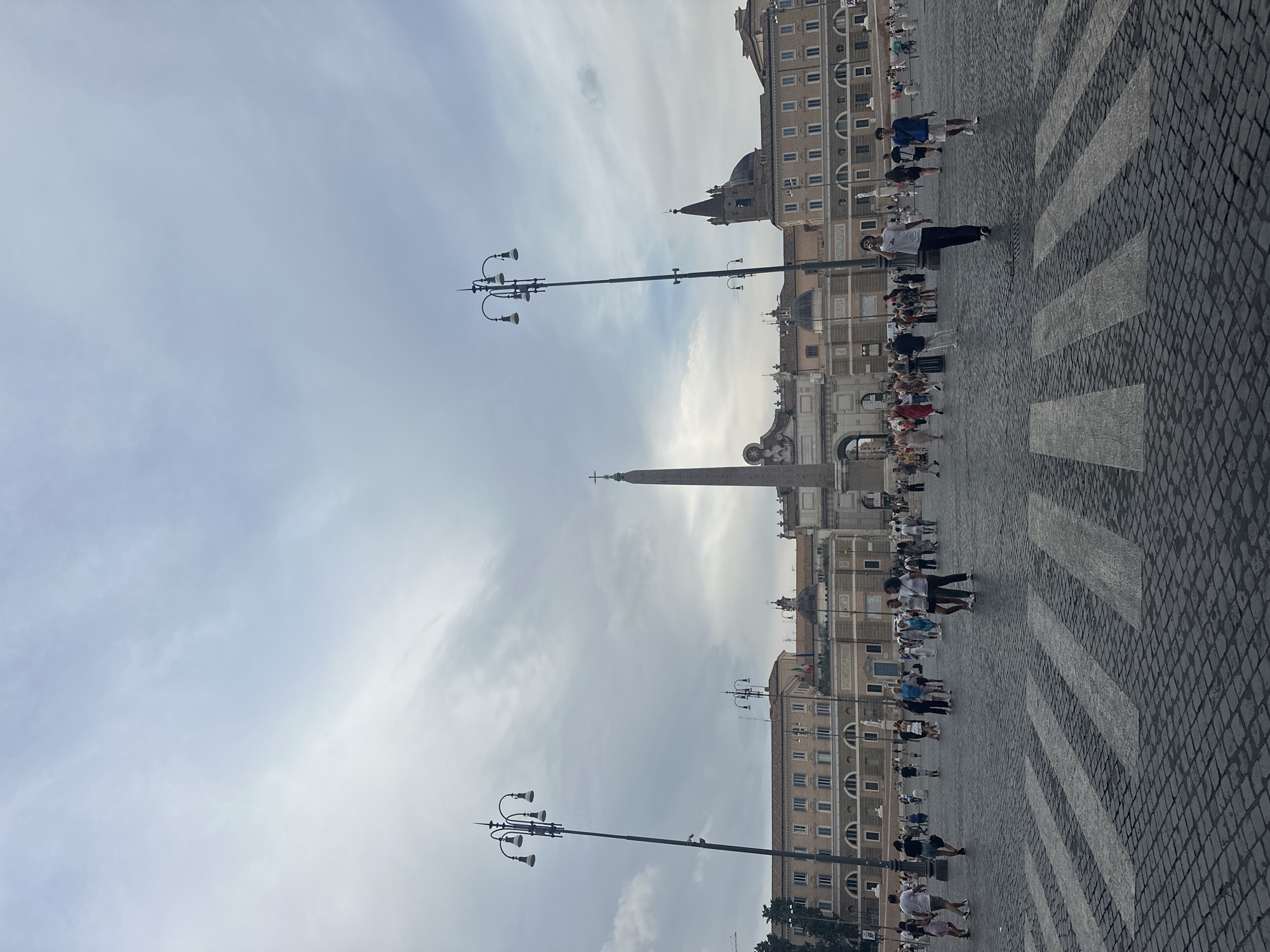The image depicts Piazza del Popolo in Rome, Italy, under a cloudy sky. A large, cobblestone plaza occupies the foreground, with a pedestrian crosswalk marked by white stripes. Numerous people are walking across the plaza in various directions. In the middle ground, an Egyptian obelisk stands tall, flanked by ornate archways and classical buildings with arched windows. Tall street lamps with multiple lights are visible on either side of the plaza. In the background, more of the city's architecture, including domes and spires, is visible. The overall scene suggests a busy public square in a historical European city.