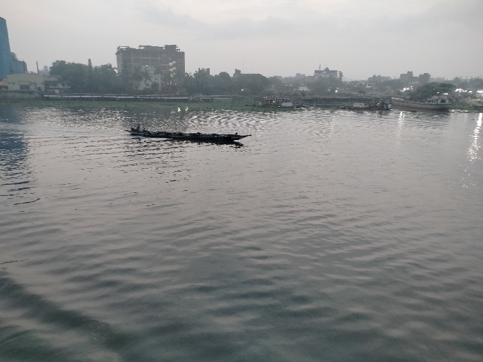 The image shows a river in Zazira, Bangladesh. It was taken from a slightly elevated position. There is a single boat with several people in it. The boat is traveling from the left of the image towards the right. The river appears calm. In the distance, there are several buildings, trees and other boats. The sky is hazy and overcast. The scene is quiet and peaceful. The location of the image is in the city of Zazira, Bangladesh, at the coordinates 23.641 latitude, 90.47 longitude.