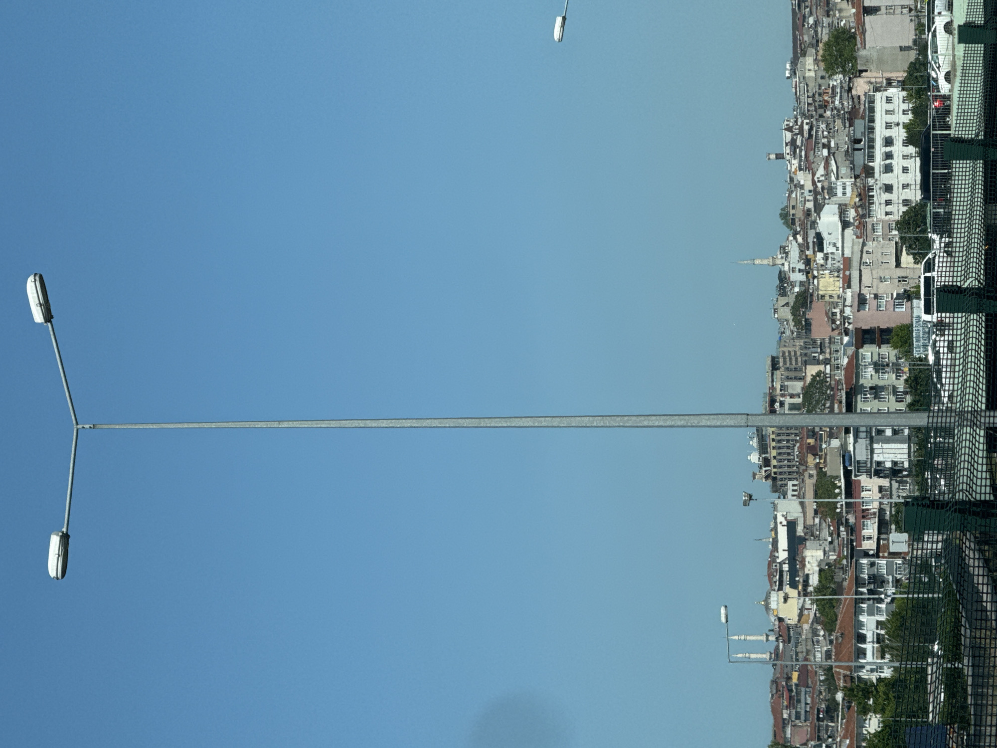 The image shows a densely populated urban area. The scene appears to be viewed from an elevated position, possibly from a building or structure. In the foreground, there's a tall metal light pole with multiple lights extending from it. The sky is clear and blue. The urban landscape consists of closely packed buildings of varying heights and designs, suggesting a historical or older section of a city. There are minarets visible, indicating the presence of mosques. The architecture includes a mix of residential and possibly commercial buildings. The buildings are predominantly light-colored, with roofs of various colors. A fence is visible in the foreground, adding a layer of depth to the composition. The location is Unknown, Unknown.