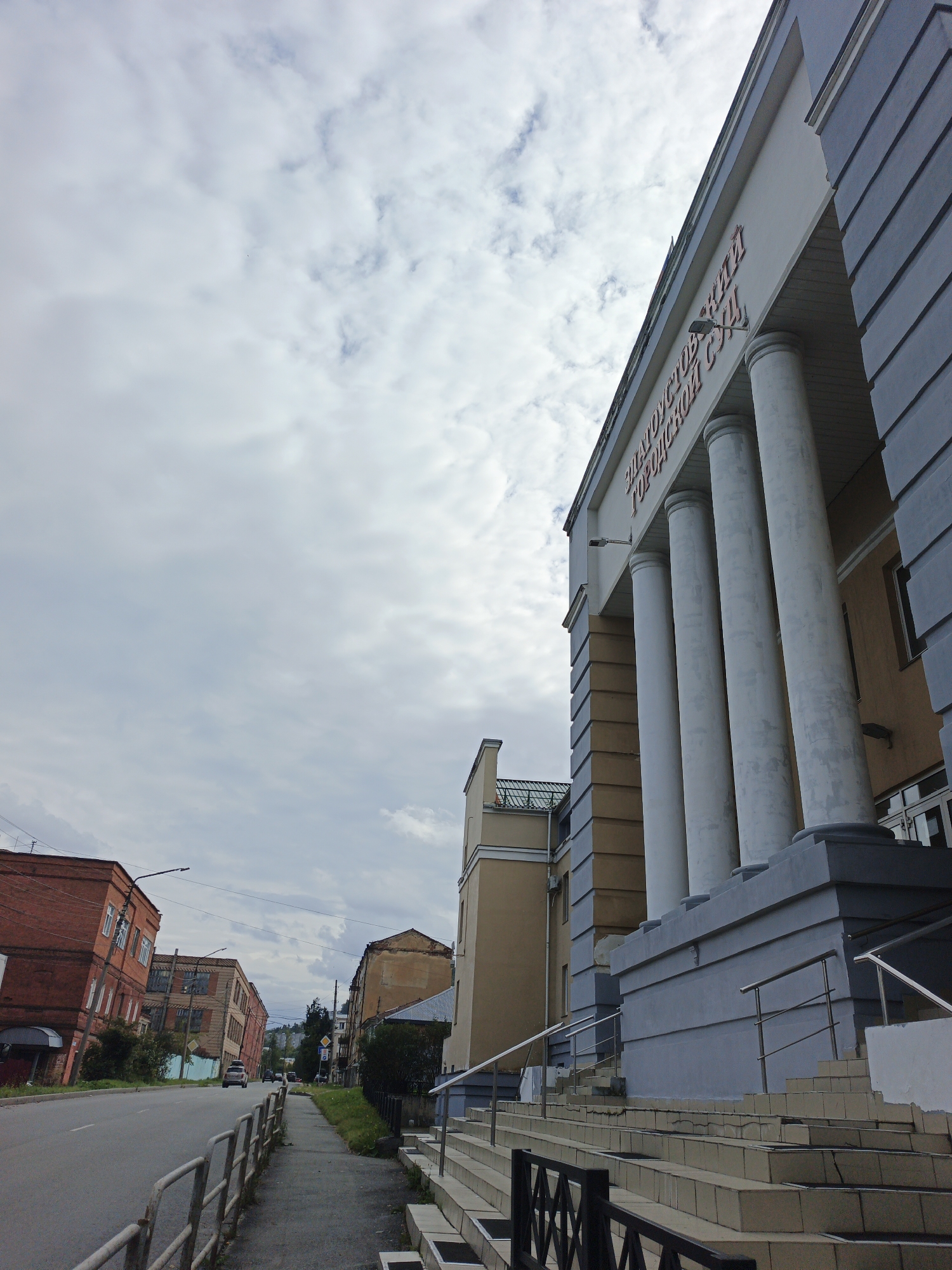 The image shows a street scene in Zlatoust, Russia, on an overcast day. In the foreground, a gray metal fence runs along the sidewalk next to a road. The sidewalk leads up to a large building with a classical facade, featuring a prominent row of white columns and red lettering that reads "ZLATOUST CITY COURT". The building has a gray base and a cream-colored upper section. To the left of this building, a street stretches into the distance, lined with several brick buildings. The leftmost brick building is red and appears to be leaning slightly. Several cars are visible on the road, with one dark SUV in the middle distance. The sky is heavily clouded with patches of lighter gray and white.