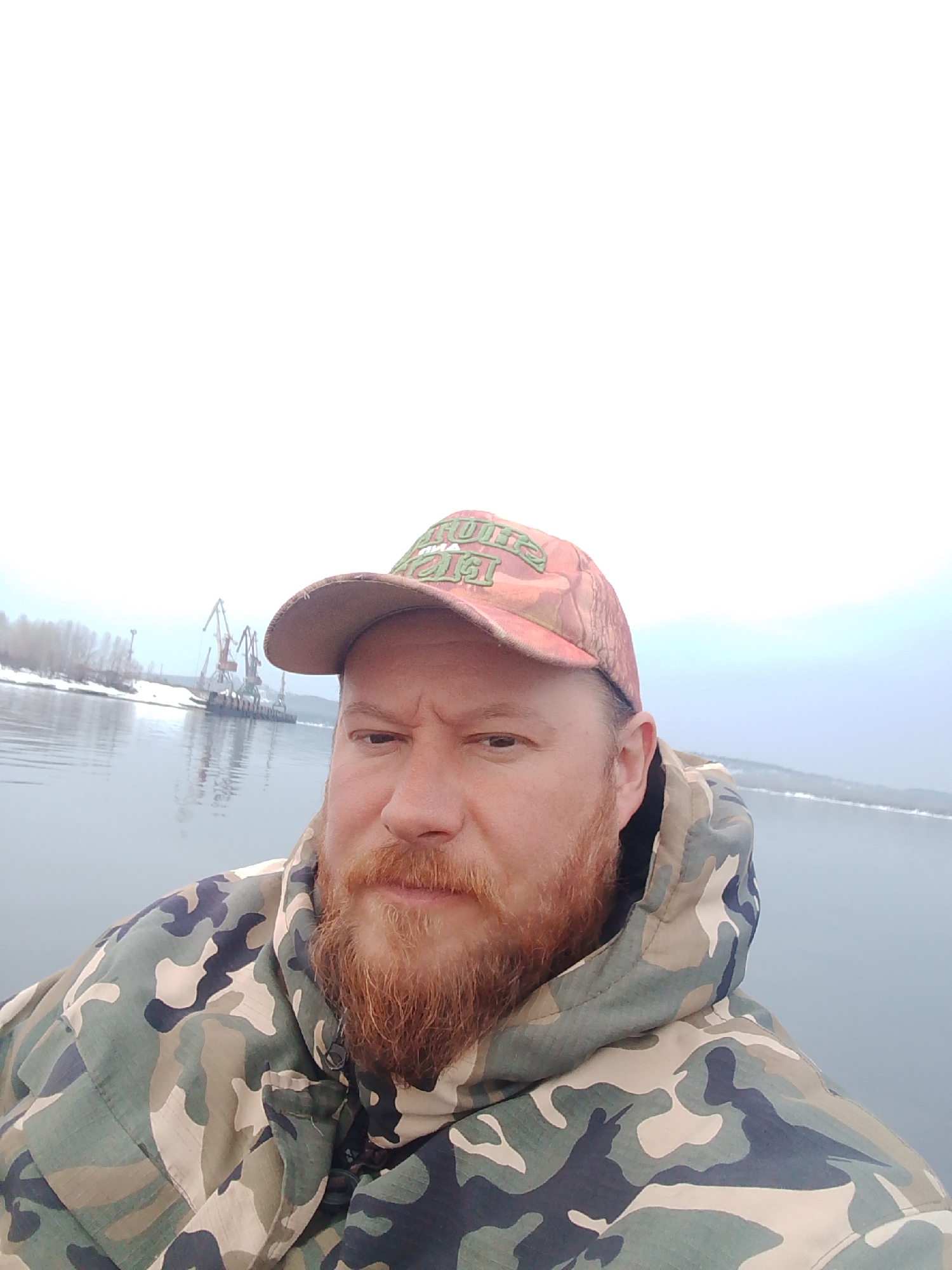 A selfie of a man with a reddish-brown beard, wearing a camouflage jacket with a hood and a camouflage baseball cap with green lettering, taken outdoors. In the background is a body of water, and on the far shore, there is snow and cranes. It is likely taken in the city of Chaikovsky, Russia.