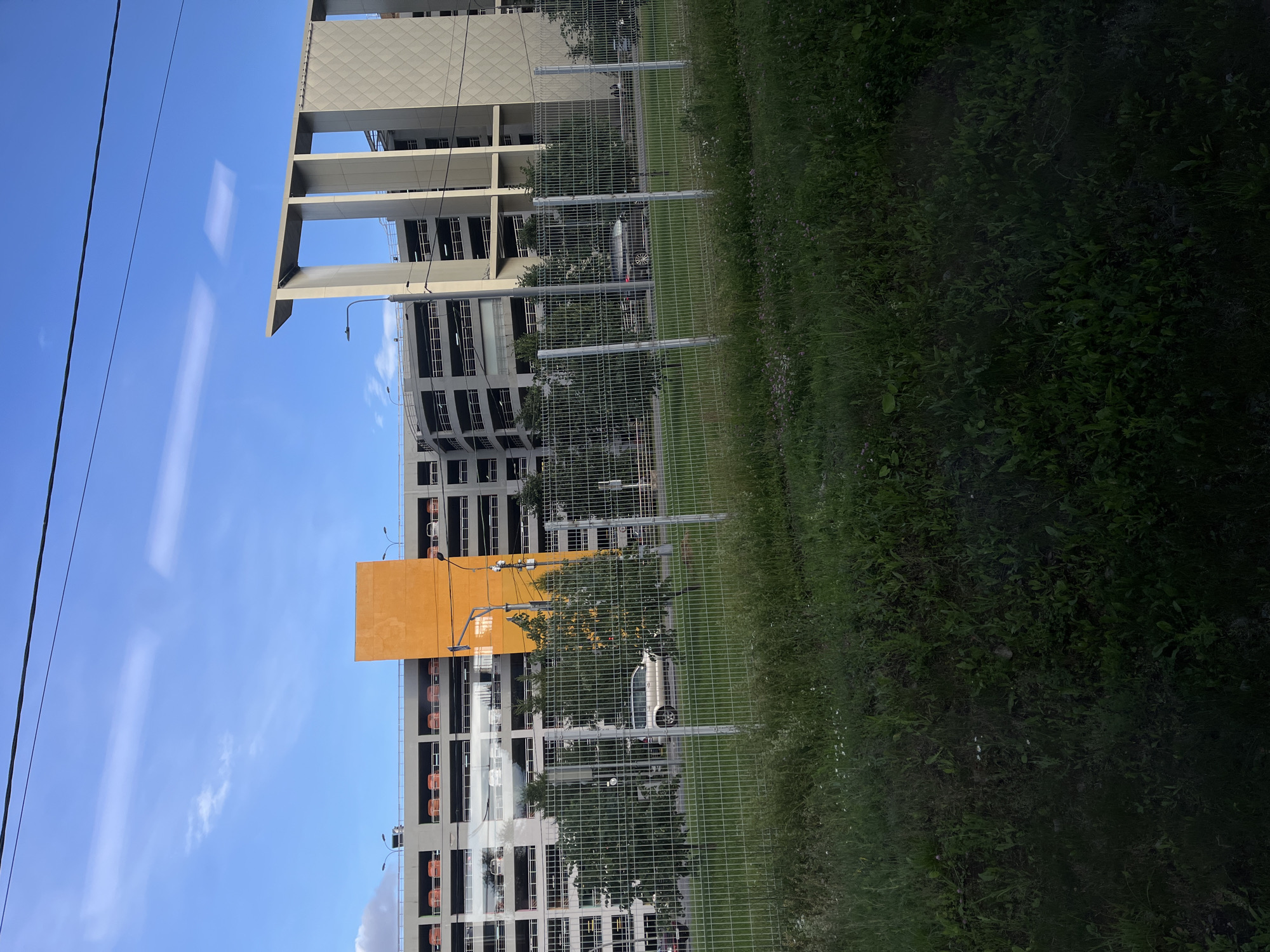 In the city of Unknown, Unknown, a large, multi-story parking garage dominates the background under a clear blue sky with wispy clouds. The garage features multiple levels with visible cars parked on each. A prominent bright orange rectangular section stands out on the left side of the structure, contrasting with the grey concrete of the rest of the building. To the right, a modern, angular section of the garage with large, spaced columns is visible. In the middle ground, a chain-link fence separates the viewer from a road where a light-colored SUV and other vehicles are driving. Trees are scattered behind the fence, and in the foreground, a patch of green grass with scattered small wildflowers is present. Overhead, power lines traverse the sky.