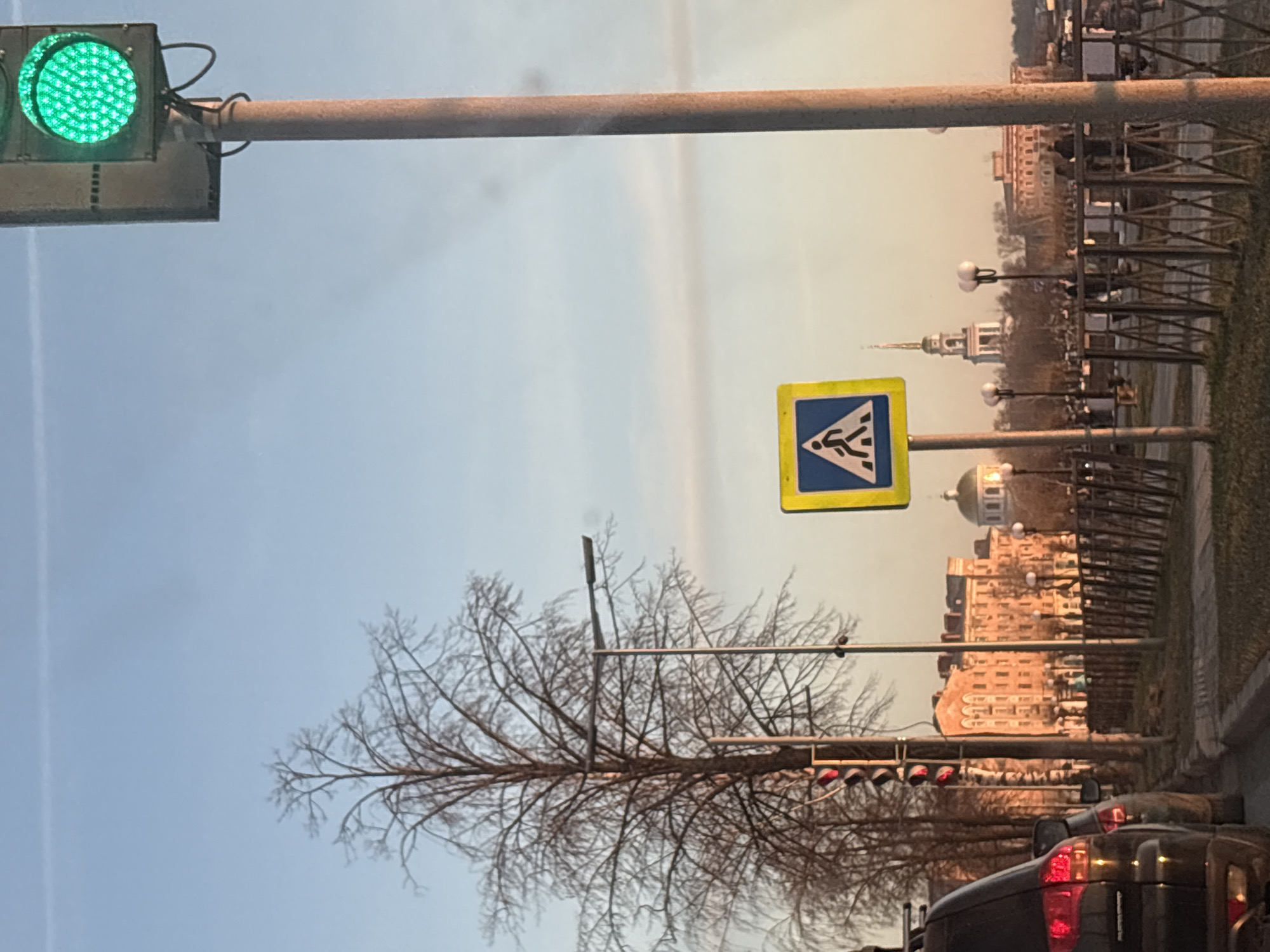 The image, when viewed upright, presents an urban street scene in Votkinsk, Russia, captured under a clear, bright sky. In the foreground, a vibrant green traffic light hangs from a metal pole, indicating an open signal for vehicles. Extending from this pole, slightly to the right, is a prominent yellow square sign featuring a blue triangle with a black stylized pedestrian figure, signifying a pedestrian crossing.
Further into the midground, a large deciduous tree with bare branches suggests a cooler season, possibly late autumn or winter. Several streetlights with spherical white globes are spaced along the roadside. The rear of a dark vehicle is visible in the lower right portion of the frame, indicating the perspective of the photo is likely from another car on the road. A second traffic light displaying red signals can be partially seen near the car.
The background reveals a row of buildings under the soft, warm glow of what appears to be late afternoon or early evening light. Notable architectural features include a building with a distinct steeple or spire and another with a traditional golden dome, common in Russian urban landscapes. A series of fences or barriers runs along the right edge of the street. The sky is a uniform, pale blue, devoid of clouds, contributing to the clear visibility of the scene. No people are explicitly visible, but the pedestrian crossing sign implies pedestrian activity in the area.