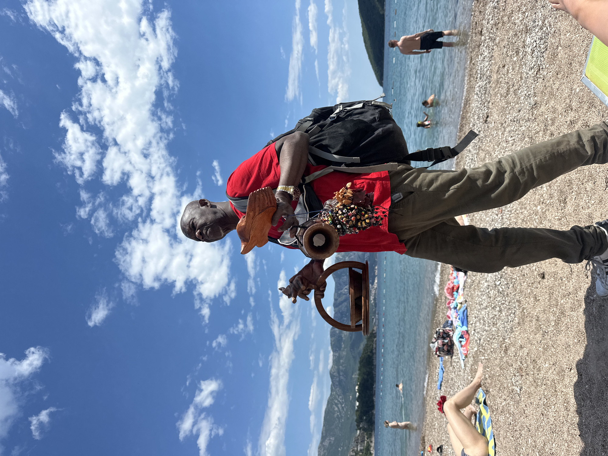 On a sunny day at a beach in Budva, Montenegro, a Black man in a red t-shirt and green pants is standing facing the camera. He has a backpack and is holding handmade crafts for sale. The beach is crowded with people sunbathing and swimming. The sky is blue with white clouds. There are mountains in the background.