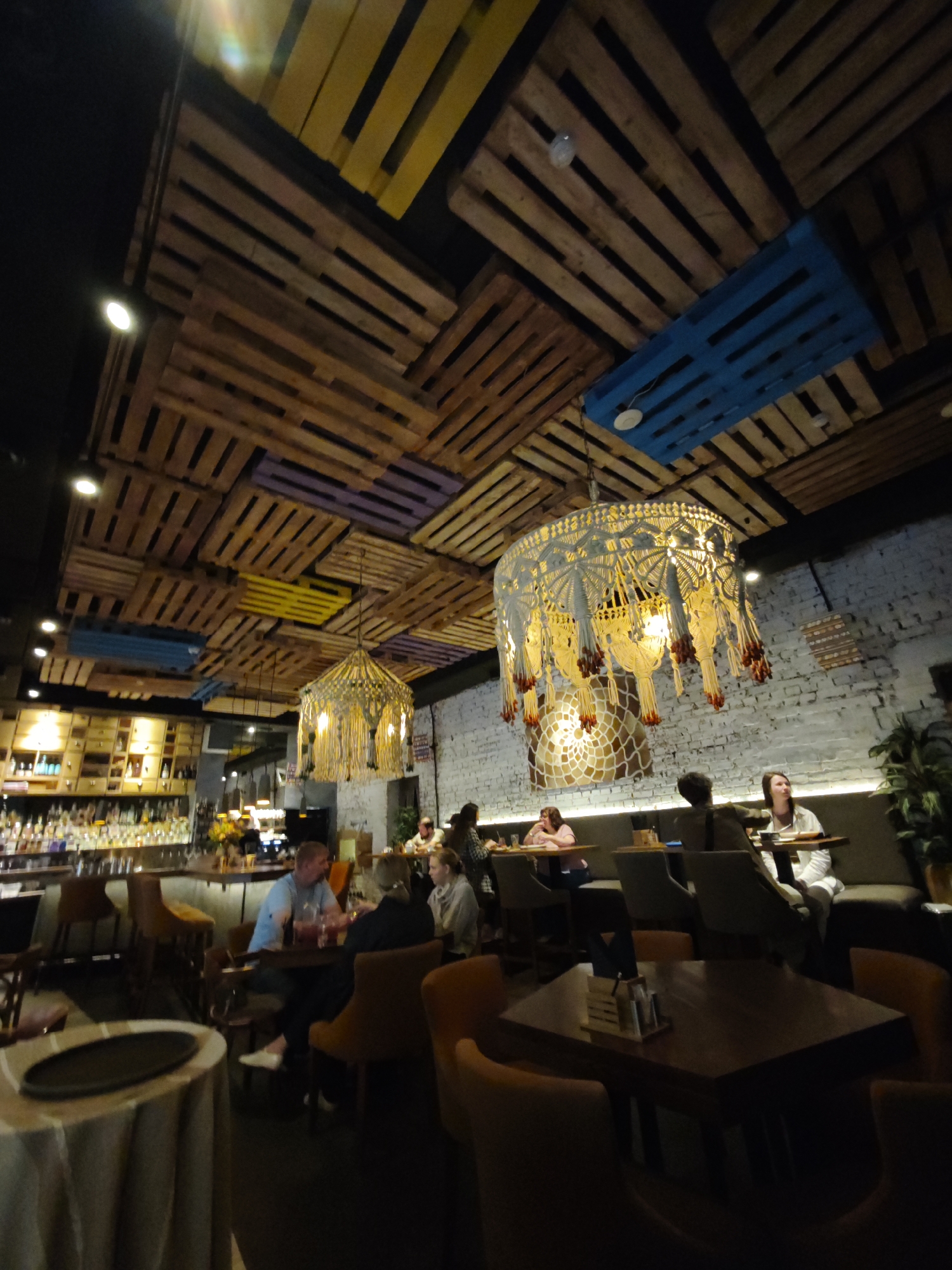 The image depicts the interior of a restaurant or bar. The ceiling is adorned with a distinctive design made of wooden shipping pallets, some of which are painted yellow, blue, and purple. Two large, intricate macrame chandeliers with hanging fringe are suspended from the pallet ceiling, illuminating the space.
In the foreground, a round table covered with a light-colored tablecloth is visible on the left. Further into the room, there are several tables with chairs. A bar area with bottles and glassware is prominent on the left side of the image.
Several patrons are seated at tables. On the right, a couple is seated at a table in the middle ground. In the background, more diners are visible at other tables. The walls appear to be made of white brick. The overall ambiance suggests a contemporary and slightly rustic dining establishment. The location is specified as the city of Saint Petersburg, Russia.