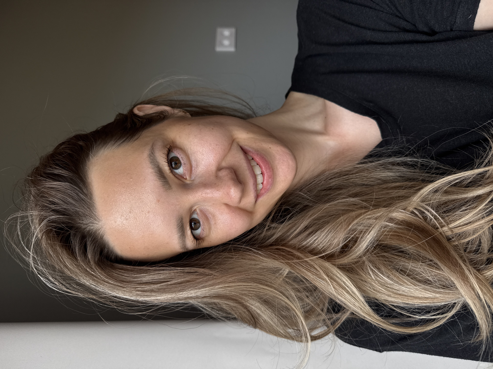 A young Caucasian woman is featured in a medium close-up, captured in an indoor setting. She is lying down or leaning back, with her head tilted and looking directly towards the camera, conveying a friendly and approachable demeanor. She has long, wavy, highlighted light brown and blonde hair, which cascades around her. Her brown eyes are open, and her eyelashes are emphasized with mascara. A clear, pleasant smile is visible on her face, revealing her teeth. She appears to have fair skin with some freckles and is wearing a plain black t-shirt or top.
The background is simple and uncluttered. Behind her, a plain dark grey wall dominates the upper portion of the frame (when the image is viewed upright). On this wall, a white electrical outlet or light switch with two receptacles is visible in the blurry distance. Below her hair and body, there is a light-colored, possibly white or off-white, surface, which could be a bed, floor, or table. The lighting is soft and even, suggesting natural light, possibly from a window, illuminating her face and hair without harsh shadows. No text is visible in the image. The scene is set in Chaikovsky, Russia.