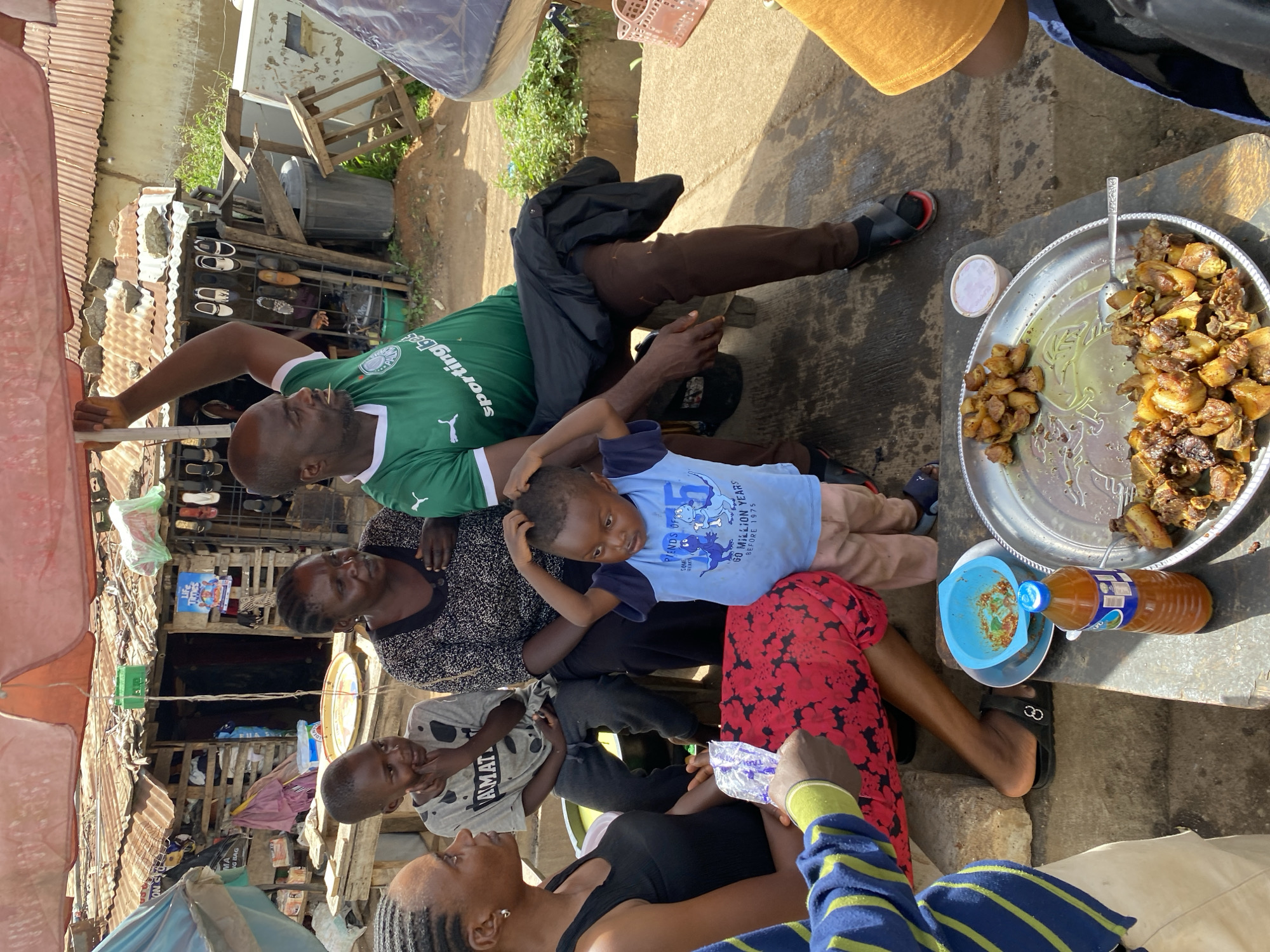 The image depicts a group of people, possibly a family, gathered around a table laden with food under a shaded area. The location is identified as the Bukuru market in Nigeria. In the foreground, a young child in a blue t-shirt and light-colored pants is being held by an adult. To their left, a woman with braided hair in a black top and red patterned skirt is seated. Behind them, a boy in a gray t-shirt and a woman in a patterned top are also seated. To the right, a man wearing a green and white soccer jersey is seated. On the table, a large platter of cooked meat, a small bowl with a reddish sauce, a bottle of beverage, and a spoon are visible. The background shows a bustling market scene with makeshift stalls, corrugated iron roofs, and various goods displayed, suggesting a typical urban market environment in Nigeria. The overall impression is one of a casual gathering where food is being shared.