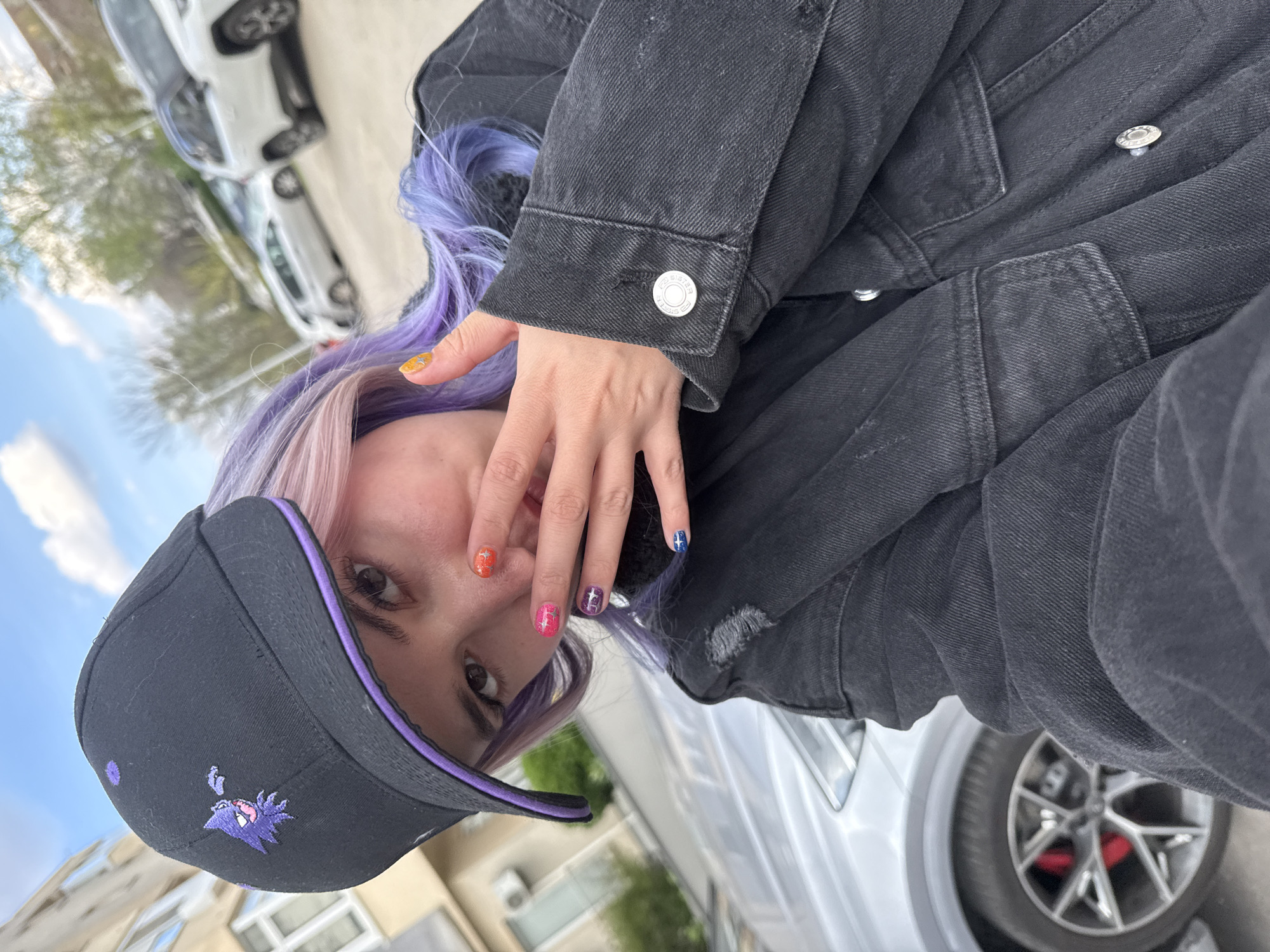 An individual, identified as a young female, is depicted in an outdoor urban environment in the City of Zagreb, Croatia. She wears a black baseball cap featuring a purple brim lining and a purple embroidered Haunter Pokémon emblem on the front. Her hair is long, displaying a color gradient from pinkish-blonde to purple. She is dressed in a black denim jacket with silver-toned buttons and visible distressed areas. Her left hand is raised, partially obscuring her mouth and lower face. Her fingernails are painted with diverse multi-colored designs, including orange with stars, pink glitter, purple-blue patterns, and dark blue with stars. She is looking directly at the camera. In the background, to the left, a light-colored building with windows and an external air conditioning unit is visible. A silver car with multi-spoke alloy wheels and prominent red brake calipers is partially visible in the foreground to the left. Additional vehicles, including a white SUV and another lighter-colored car, are out of focus in the background. The sky above is blue with scattered white clouds, indicating daytime conditions.