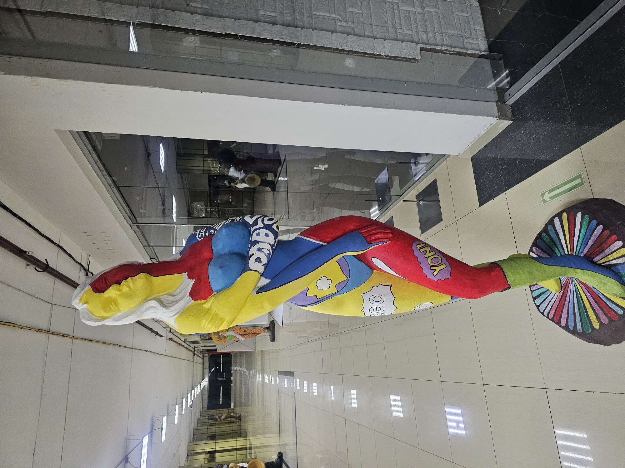 In the city of Nakhodka, Russia, a colorful, abstract sculpture of a female figure stands prominently in an indoor public space, likely a mall or gallery, with a polished, tiled floor reflecting overhead fluorescent lighting. The sculpture is a vibrant display of primary colors and cartoonish elements, with one side of the figure's face painted yellow and the other red, separated by a white stripe. The body is a patchwork of red, blue, and yellow, featuring comic-book-style speech bubbles with text such as "ec" and "YON!" The sculpture's base is a circular platform adorned with a radial pattern of rainbow colors. In the background, the hallway extends into the distance, with other sculptures and figures visible, as well as storefronts with glass facades.