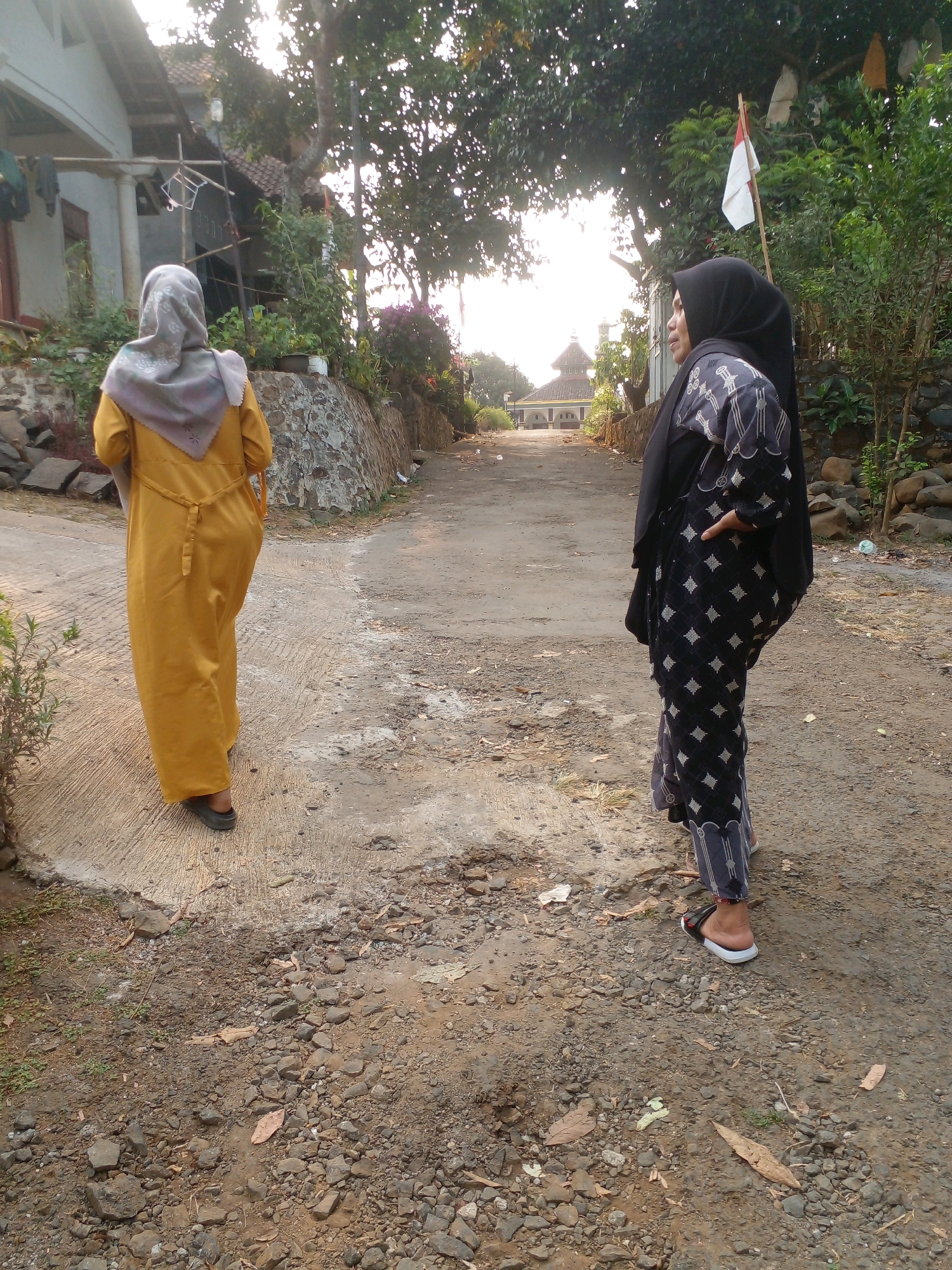 In the city of Pungangan, Indonesia, a dirt road meanders through a rural landscape, flanked by stone walls and lush vegetation. The scene is illuminated by the soft glow of a setting sun, casting long shadows and highlighting the earthy tones of the surroundings.
Two women, dressed in traditional Islamic attire, stand on the road. The woman on the left wears a flowing mustard-yellow dress and a grey headscarf, her back to the viewer. The woman on the right, facing left, is clad in a black hijab and a patterned black and white dress, her hand resting on her hip.
In the background, a mosque with a red-tiled roof and a white minaret stands in the distance, a testament to the community's spiritual heart. A red and white flag, the national flag of Indonesia, flutters gently from a wooden pole, adding a touch of patriotism to the tranquil scene. The overall impression is one of peace and serenity, a snapshot of daily life in a remote Indonesian village.