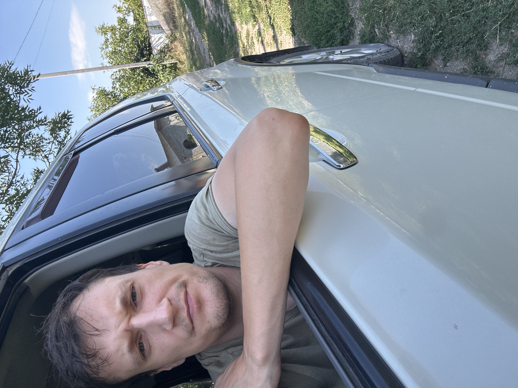 A man is leaning out of a light-colored SUV window. He has short, dark hair and is wearing a green t-shirt. His left arm is resting on the edge of the open window. The vehicle appears to be parked on a dirt road, with grass and trees on either side. A white fence and a utility pole are visible in the background. The sky is clear and blue. The man is looking directly at the camera with a neutral expression. The location is Makiv, Ukraine.