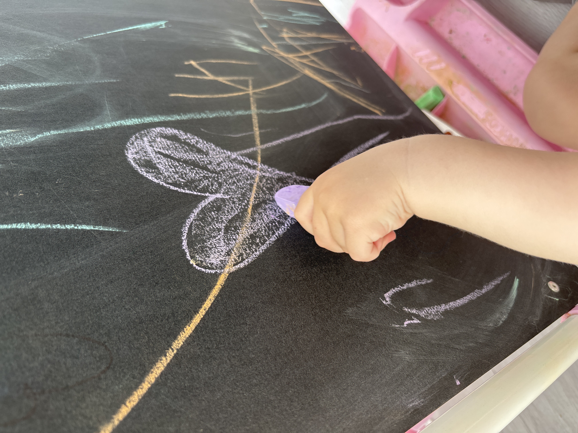In an indoor setting, an unseen child's hand is holding a piece of purple chalk, drawing a heart shape on a black chalkboard. The chalkboard has other colorful chalk markings, including a yellow diagonal line and some vertical strokes in green and orange. A pink plastic tray with some green chalk and smudges is visible in the lower right corner of the image, suggesting an art activity. The background is out of focus.