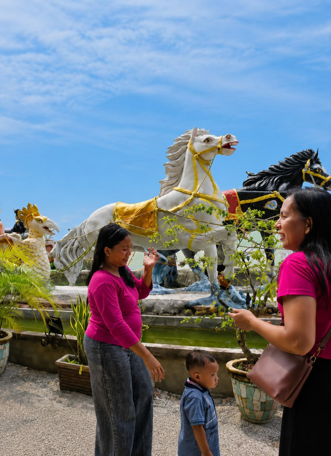The image depicts an outdoor scene at what appears to be a tourist attraction in Kota Pari, Indonesia. In the foreground, two women in vibrant pink tops are interacting, one looking towards the other and gesturing with her hand. A young child in a blue shirt stands between them, looking off to the side. Behind the people are large, ornate statues of horses, one white with a golden saddle and harness, and another black. To the left, a statue of a mythical creature resembling a bird with a crown is visible. The statues are positioned near a body of water, likely a pond or pool, surrounded by potted plants. The background features a clear blue sky with scattered clouds. The ground is covered in gravel or small stones. The overall atmosphere suggests a sunny day at a decorative outdoor location.