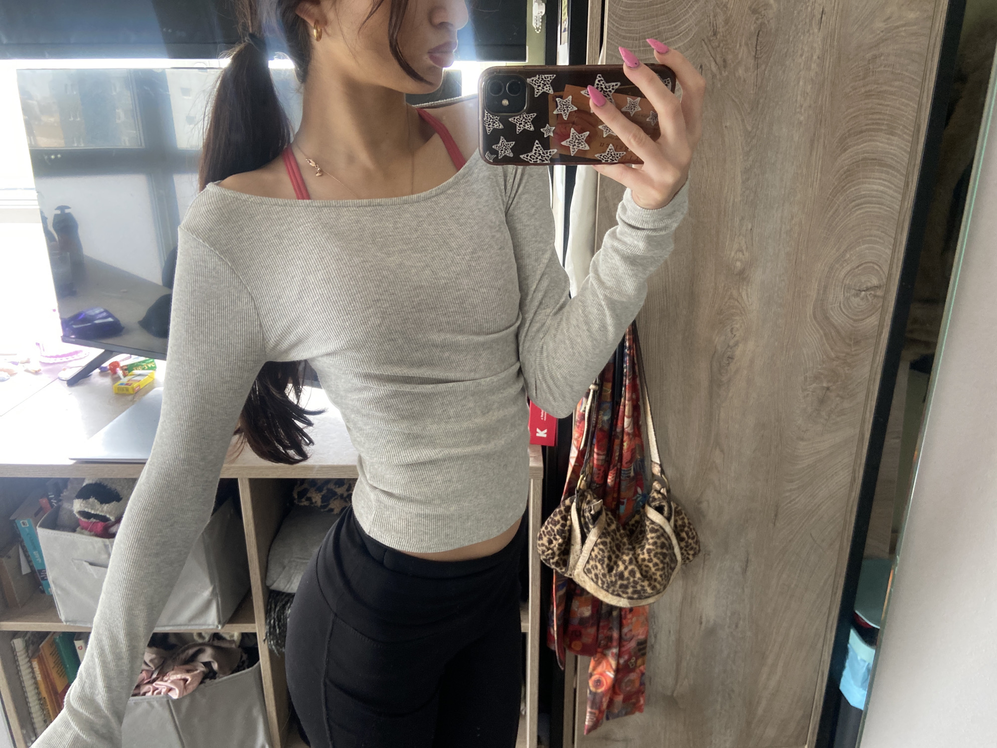 A person takes a mirror selfie. The person is wearing a gray long-sleeved shirt, black pants, and a gold earring, with their hair pulled back in a ponytail. The phone case is black and decorated with white stars. The background includes a bookshelf with bins, a scarf and leopard print purse hanging on a wooden closet door, and a mirror reflecting a window and other items. The location is not identifiable as London.