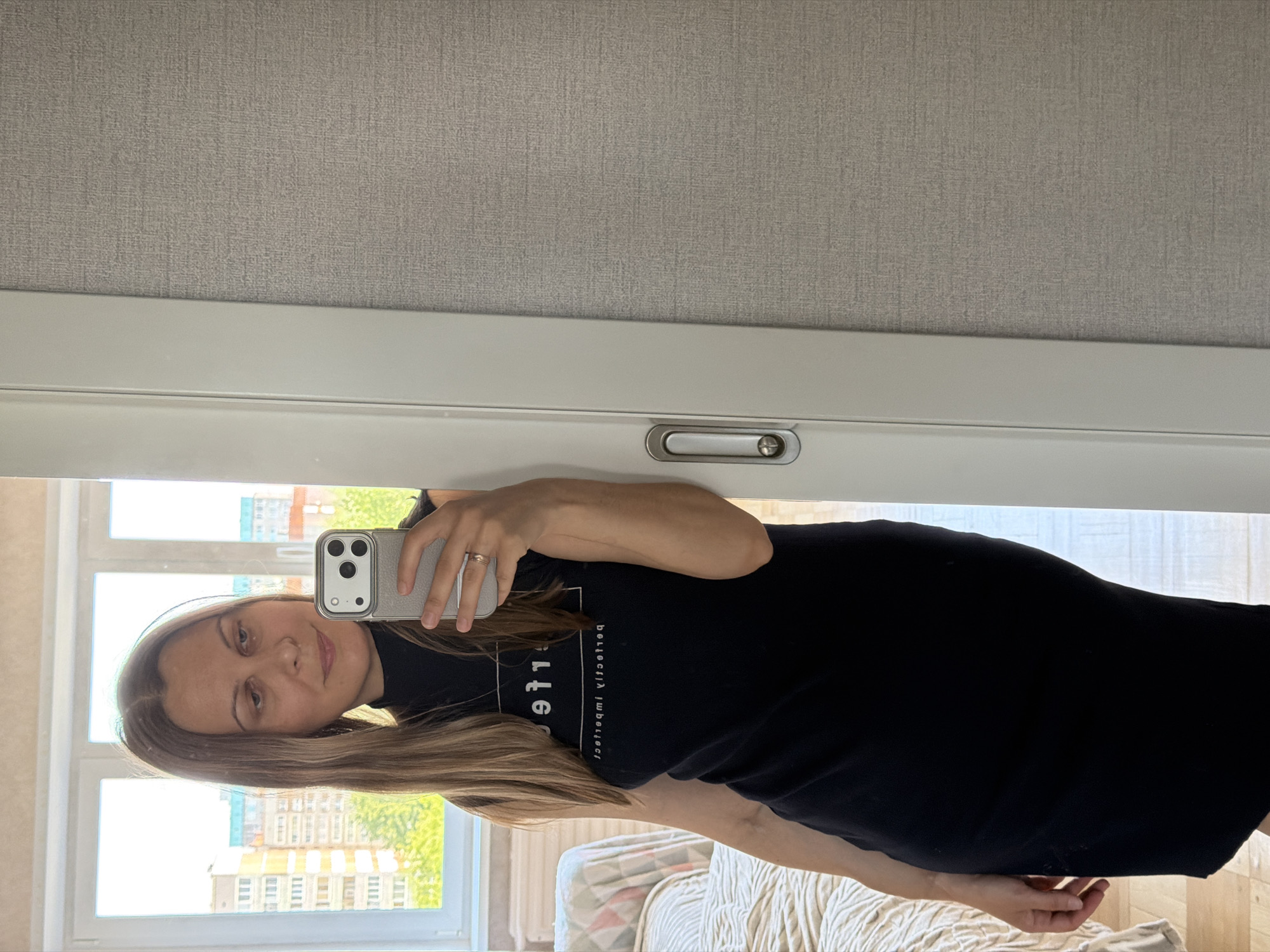 A woman with blonde hair, wearing a black sleeveless dress with white lettering and a ring on her left hand, stands by a white doorway. She is holding a silver smartphone in her right hand, taking a mirror selfie. The background reveals a window with a view of apartment buildings and trees, suggesting an urban setting. To the right of the woman, a textured gray wall is visible. The scene appears to be indoors, likely in a residential apartment in Izhevsk, Russia, given the presence of residential buildings visible through the window.