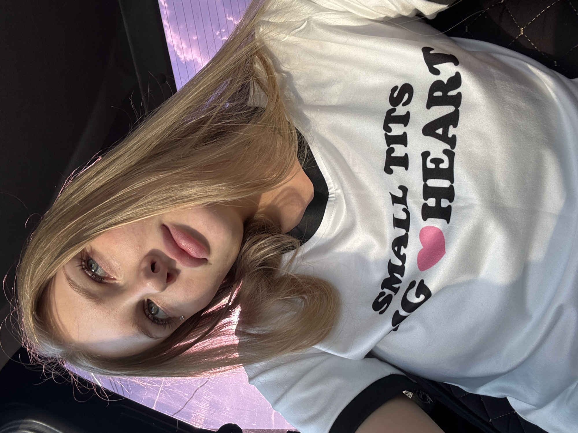 A young woman with blonde hair and blue eyes is photographed from the chest up, looking towards the viewer's left. She is wearing a white t-shirt with black trim around the collar and sleeves. The t-shirt has black text and a pink heart graphic on it. The text reads "SMALL TITS" on the top line and "BIG HEART" on the bottom line, with the pink heart replacing the letter "O" in "BIG". The background shows a purple-tinged sky with clouds, possibly through a car window. The lighting suggests it is daytime, with sunlight illuminating the woman's face and hair. No specific location in the city of Perm, Russia can be identified from the image.