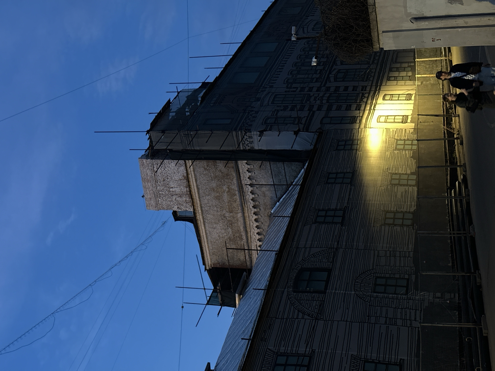 Here is a description of the image:
The image depicts a tall building in Moscow, Russia. The facade of the building is mostly covered in a grey material that is patterned with black lines in the style of an architectural drawing that depicts the facade of the building itself. Scaffolding is present along the building, and what appears to be a tarp is used as a covering on some parts of the building. There is a protruding brick structure in the upper portion of the image. Overhead wires are visible against a blue sky in the upper left. In the lower right are two people walking on a street or sidewalk. A street lamp is on.
