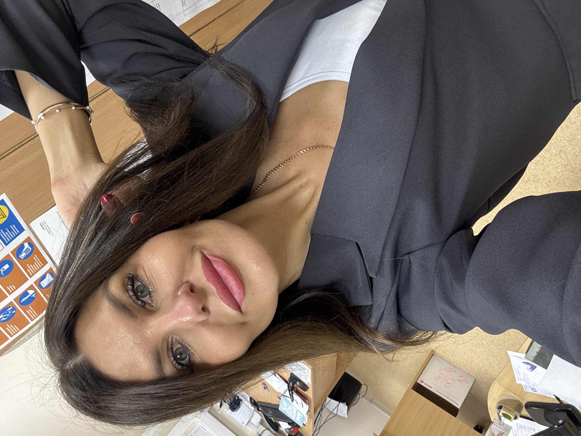 A medium shot, slightly angled selfie of a woman in an office. The woman has long, dark brown hair, green eyes, and is wearing a dark gray blazer over a white top. She has on pink lipstick and her left hand is raised behind her head, showing a pearl and gold bracelet. The background is an office setting with a wooden desk visible, littered with papers, a computer keyboard, and other office supplies. A corkboard with safety signage is mounted on the wall behind her. The overall lighting is bright, suggesting an indoor environment.