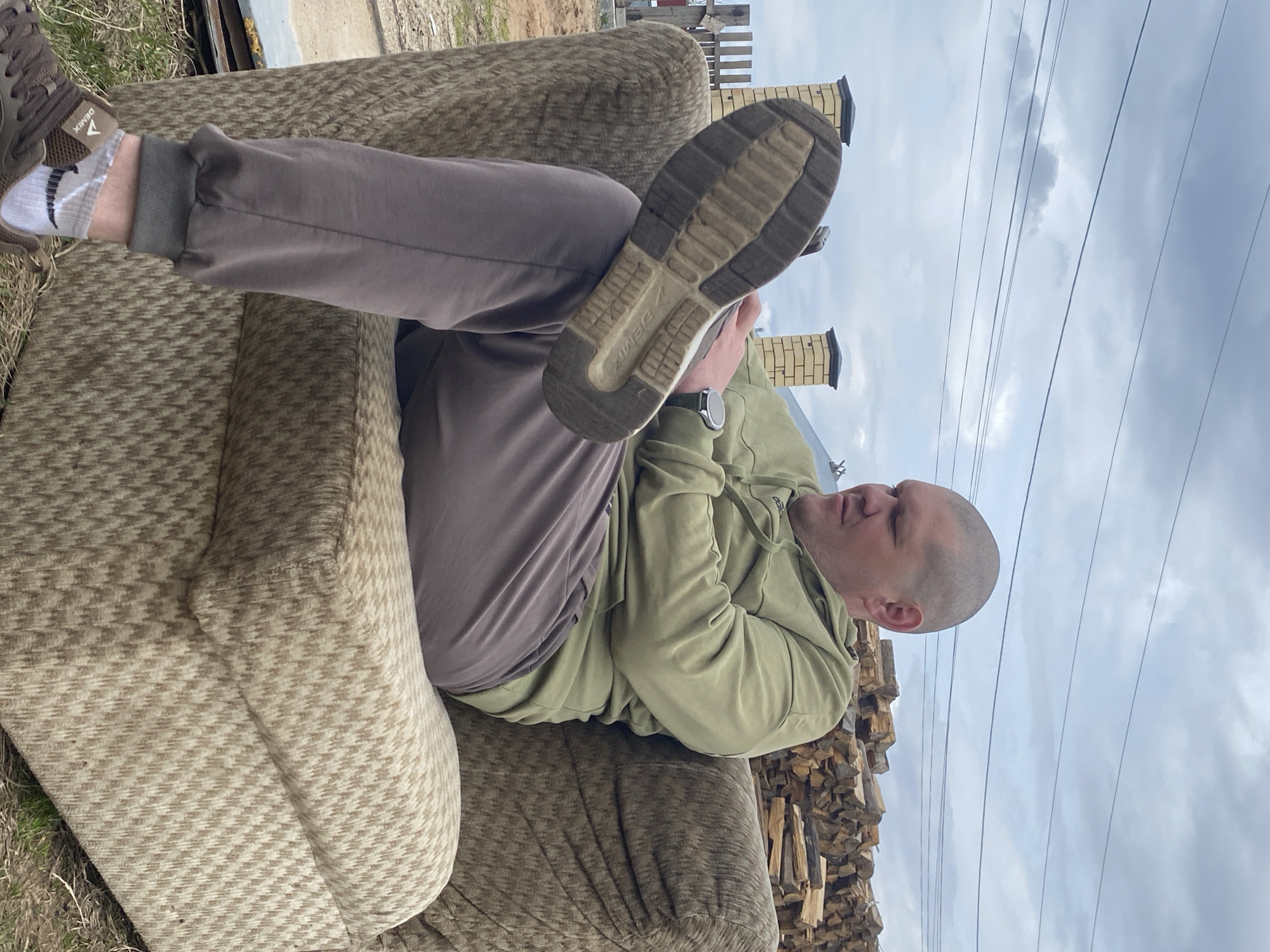 A man is reclining on a worn, patterned couch outdoors. He is wearing brown sweatpants, a light green hooded sweatshirt, a wristwatch, and white socks with gray accents. His right leg is elevated, displaying the sole of a Demix sneaker toward the viewer. The man has a shaved head and appears to be looking at the shoe on his foot. Behind the couch is a stacked pile of firewood. In the background, there's a building with two brick chimneys and a cloudy sky. The location is likely Chaikovsky, Russia.