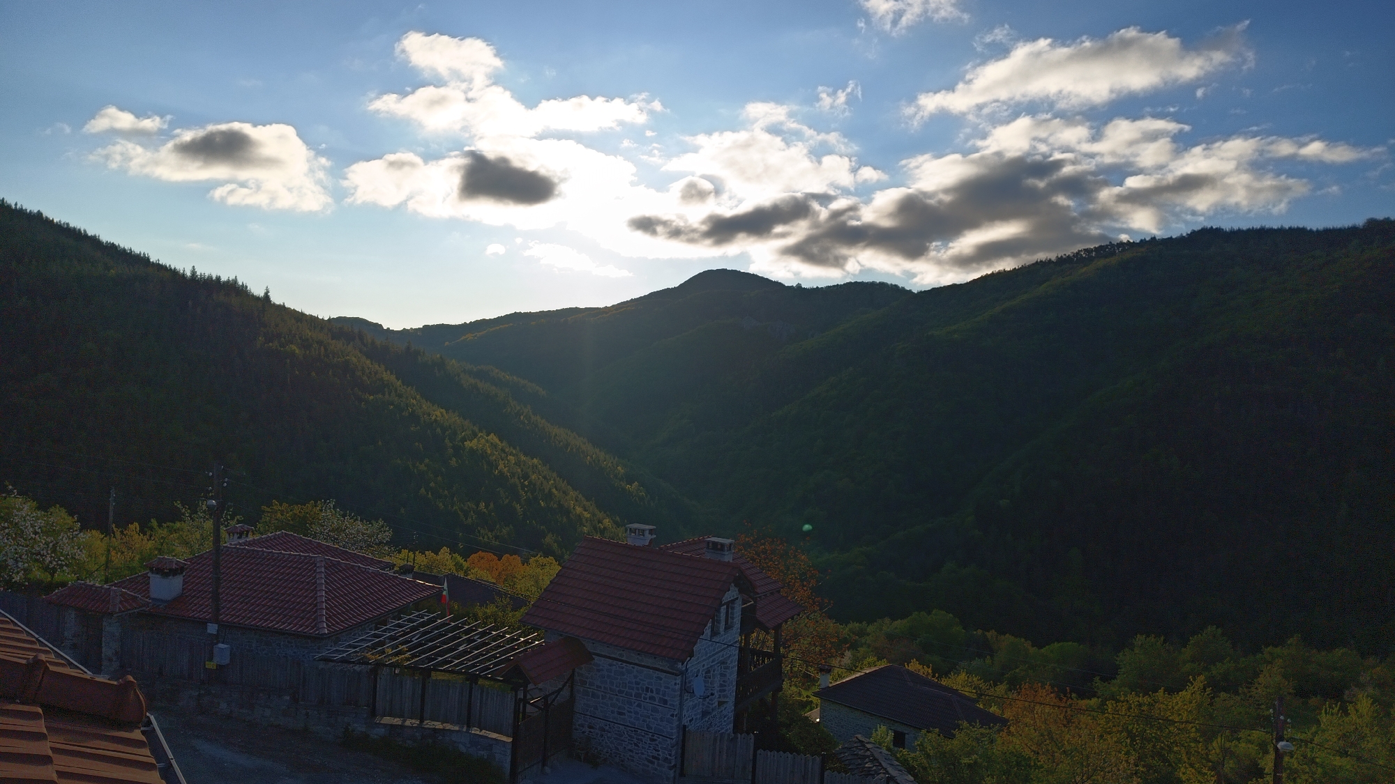 The image shows a landscape in Vrata, Bulgaria, featuring mountains, houses, and sky. In the foreground, there are several houses with red tile roofs, surrounded by trees. The middle ground contains a valley between the mountains, which are covered with dense vegetation. The sky is blue with scattered white and gray clouds. The sun is obscured by the clouds, creating a diffused lighting effect.