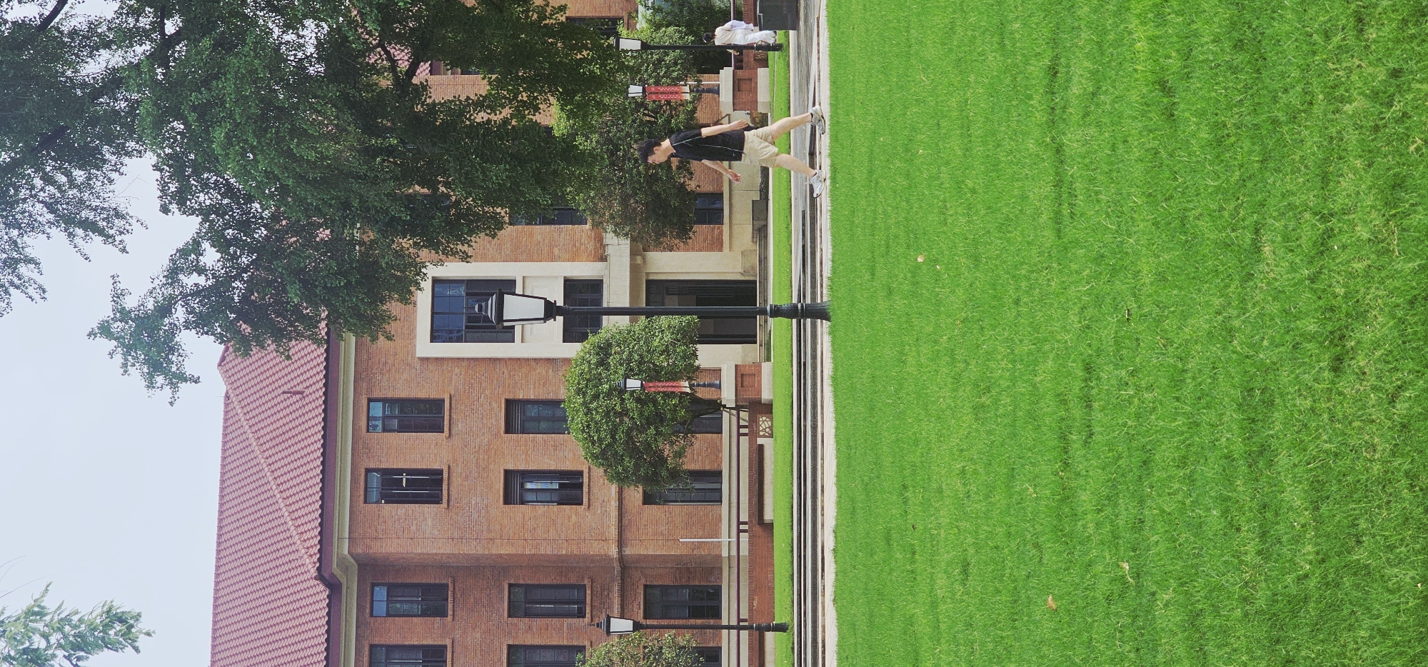 A medium shot depicts a person jumping in the air near a building in the Yuzhong District, China. The individual is dressed in dark-colored attire, complemented by lighter-toned shorts and athletic shoes. They appear to be in mid-leap, oriented towards the right side of the frame.
The backdrop consists of a multi-story brick building with a reddish-tiled roof, featuring multiple windows and what appears to be a centrally positioned entrance. Lush green lawns are visible. A few lampposts with ornate bases stand on the grass near the building. Green trees are interspersed, adding to the natural setting surrounding the building.