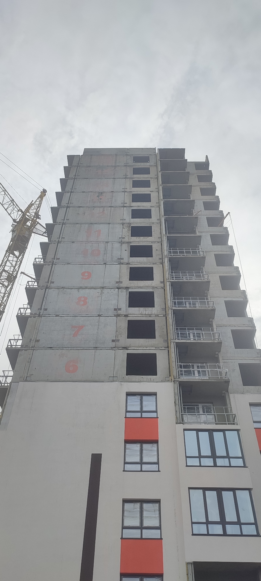 The image shows a multi-story building under construction in Novoaltaysk, Russia. The building is made of concrete and has a crane on the left side. The windows are either dark or covered with protective plastic. Some of the concrete panels on the exterior of the building are marked with red numbers, indicating floors or sections. The sky is overcast, suggesting a cloudy day. The lower portion of the building appears to be more finished, with some windows installed and sections of orange facade.