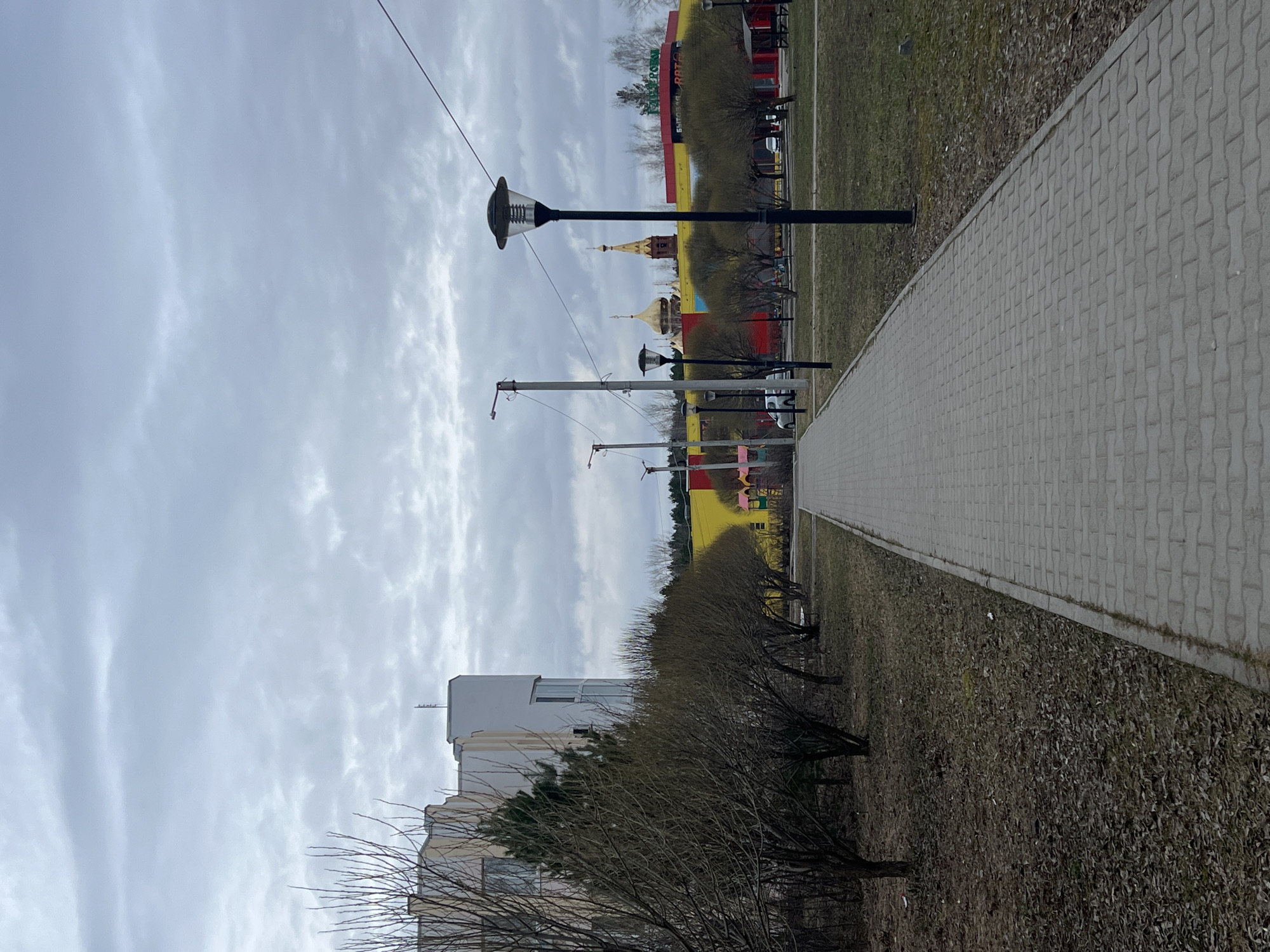 A sidewalk runs along a green space in Chaikovsky, Russia. There are lampposts alongside the sidewalk. A building is on the left and a structure with gold domes and bright colors in the background. The sky is cloudy.