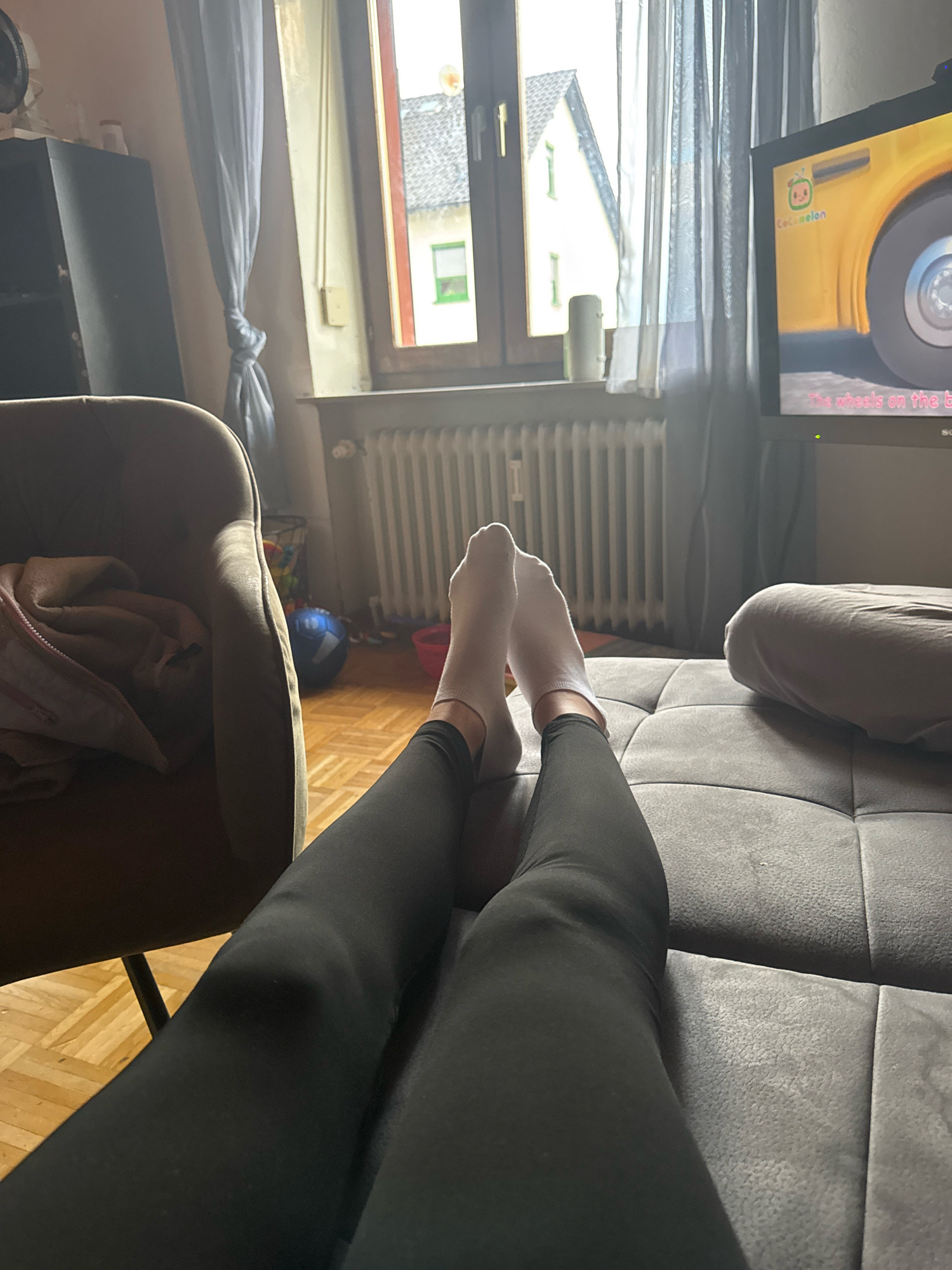 This image depicts a person's legs and feet, clad in white socks and black leggings, resting on a grey tufted couch. The perspective is from a low angle, looking up towards the television screen which is playing "CoComelon," displaying animated characters and lyrics. To the left of the couch is a brown armchair with a folded blanket. In the background, a window reveals a street view of houses in Überherrn, Germany, with a radiator below the window. The overall ambiance is relaxed and domestic, suggesting a moment of leisure.