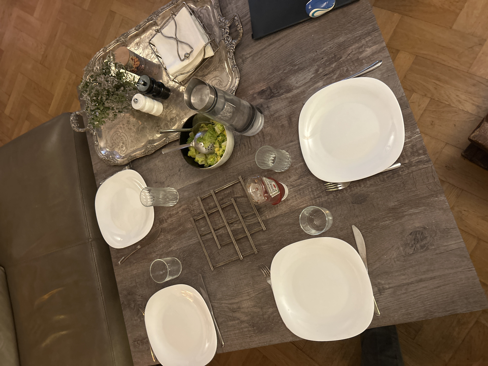 The image shows a table set for three people, possibly in a home setting. Three white plates are arranged, each with a fork, knife, and glass. A silver tray sits on the table, holding a salad bowl, water pitcher, salt and pepper shakers, a small bouquet of flowers, and napkins. A metal rack and a bottle of ketchup are placed near the center of the table. The table has a dark, wood-like surface and is next to a leather couch. The room appears to be indoors, with parquet flooring visible in the background.
