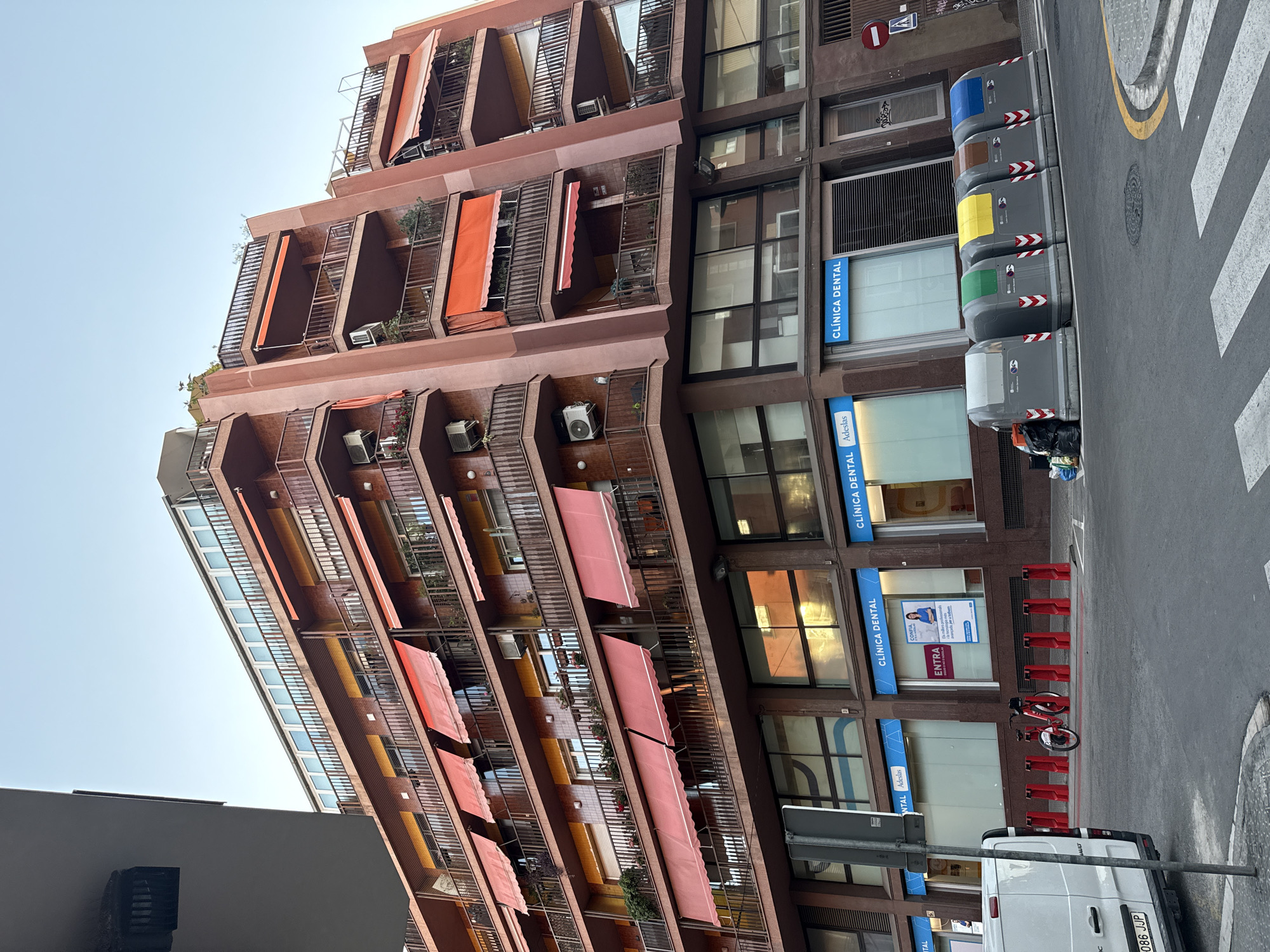 The image captures a street-level view of a multi-story building in Barcelona, Spain, on a clear day. The building features a distinctive pinkish-brown facade with multiple balconies, each equipped with reddish-orange awnings. Several air conditioning units are visible on the balconies. The ground floor of the building houses commercial spaces, with prominent signage indicating "CLÍNICA DENTAL" (Dental Clinic) in blue lettering.
In front of the dental clinics, there is a row of red bollards, and behind them, two red bicycles are parked. To the left, the rear of a white van is visible, displaying a license plate ending in "JP". Further to the right, a line of five large, grey recycling bins are arranged, each with a different colored lid and chevron markings. These bins are filled with refuse. Adjacent to the bins, there are various pieces of garbage, including black trash bags.
The street itself is paved, with a yellow and white striped pedestrian crossing visible in the foreground. The sky is a clear, pale blue. The architecture of the building, particularly the design of the balconies and awnings, is characteristic of urban European cities. The presence of the recycling bins and the parked bicycles suggests a public space within a city environment.