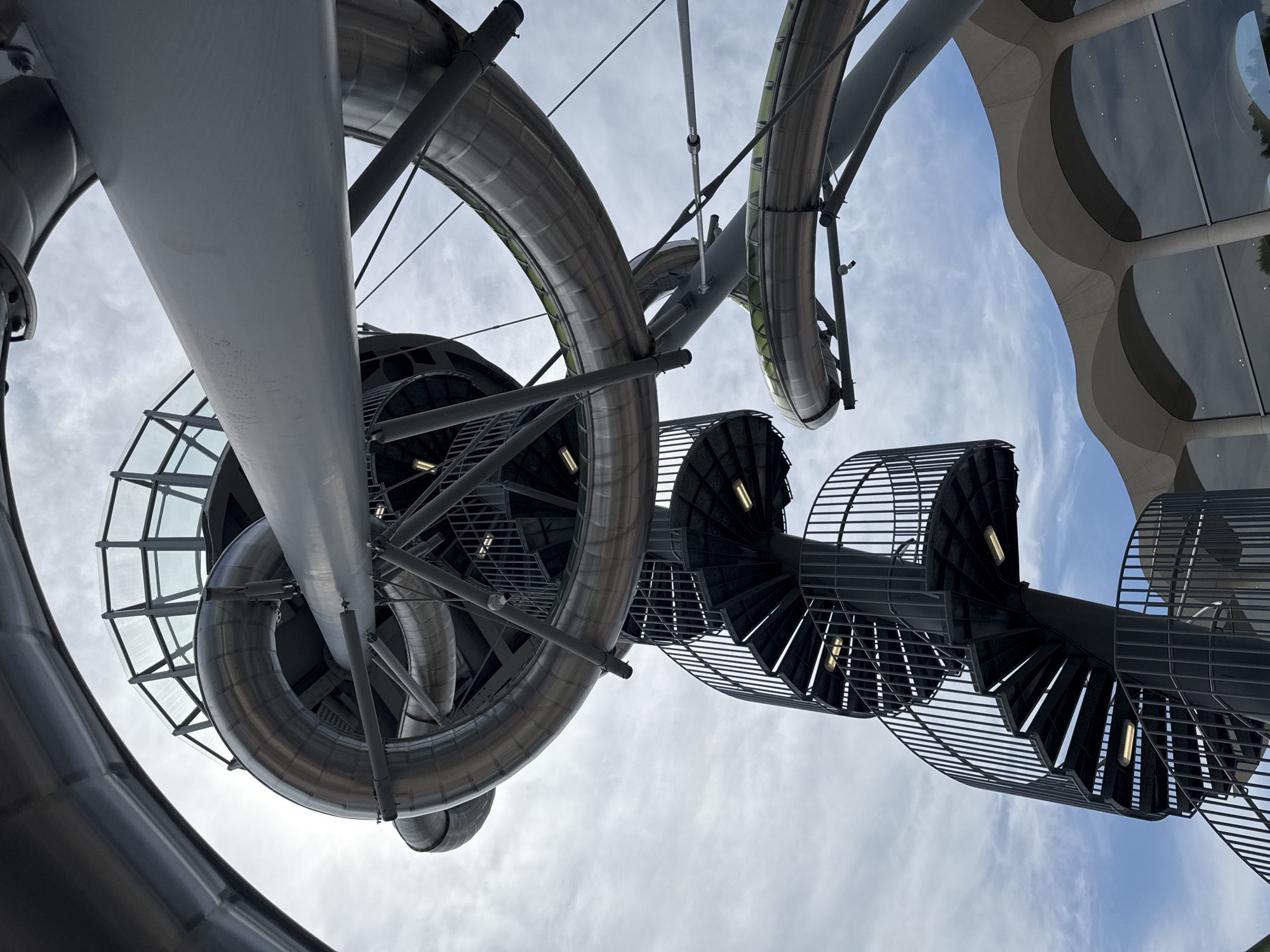 The image captures a low-angle perspective of a complex spiral waterslide structure against a cloudy sky, likely located in Aventura, Florida. The foreground features a dark metal spiral staircase with railings, illuminated by several lights along its ascending path. Above and behind the staircase, a large, silver, tubular waterslide snakes and curves through the frame. Parts of the waterslide appear to have transparent green sections. A glass-enclosed observation area is visible at the top of a section of the waterslide. In the background, a portion of a building with a distinctive wavy, light-colored roofline and large, reflective windows can be seen. The overall impression is of a modern, architectural attraction.