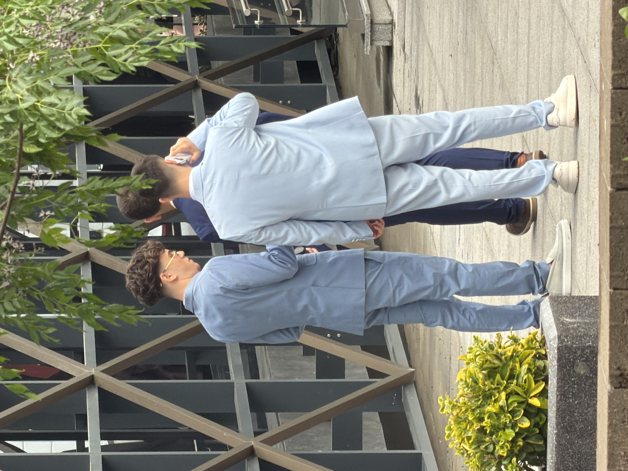 Two men are standing outside, likely in the city of Burgas, Bulgaria. One man is holding a phone near his head, possibly talking on it. Both men are wearing light blue suits, suggesting they might be attending an event or gathering. The background includes a modern building with geometric metal detailing and some greenery. One man wears white sneakers and the other wears brown dress shoes.