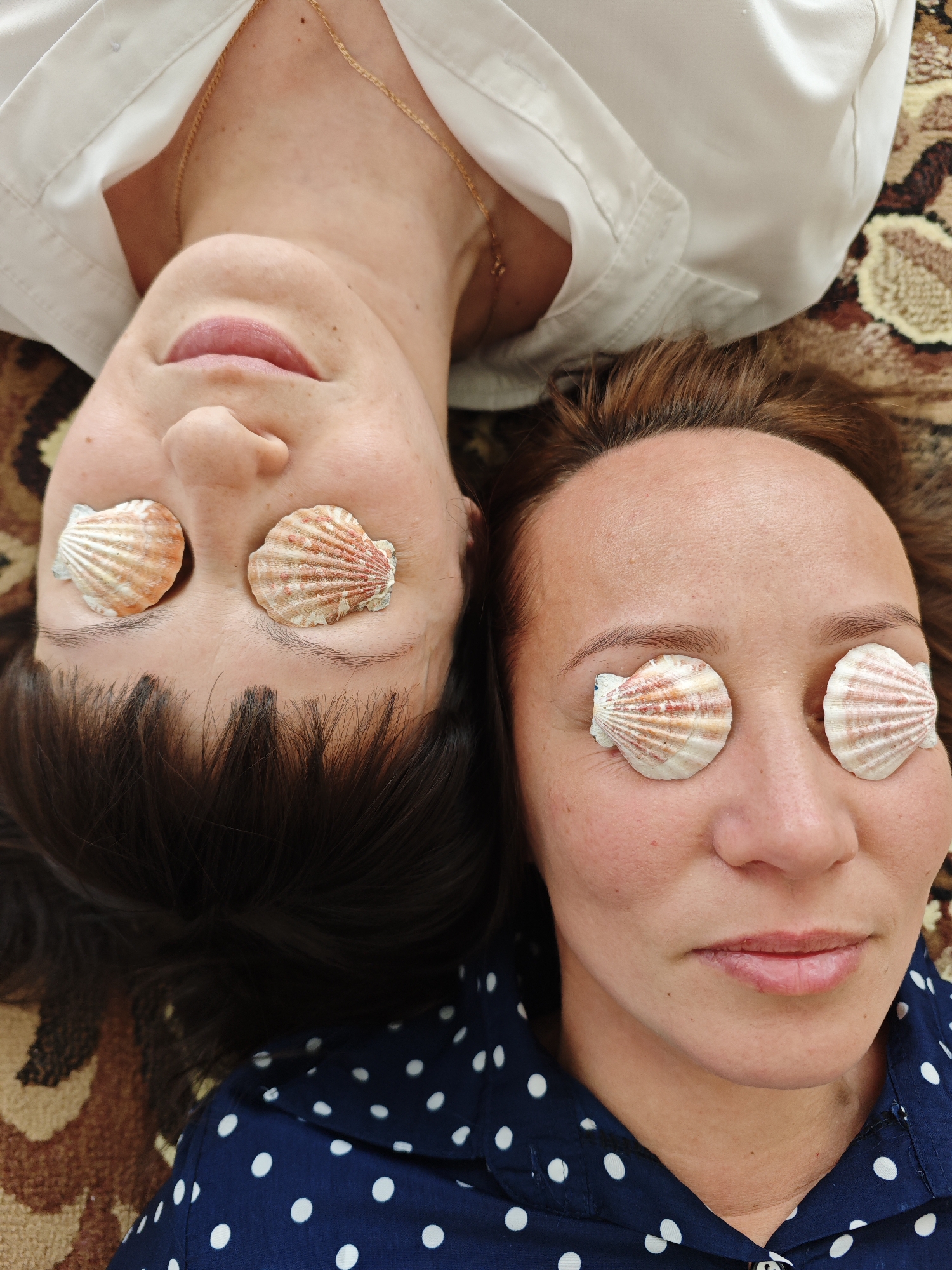 Two women are lying head-to-head on a carpet. Each woman has a seashell covering each eye. The woman on the left is wearing a white collared shirt and has dark hair. The woman on the right is wearing a navy blue and white polka dot shirt and has reddish-brown hair. The location is Markovskii, Russia.